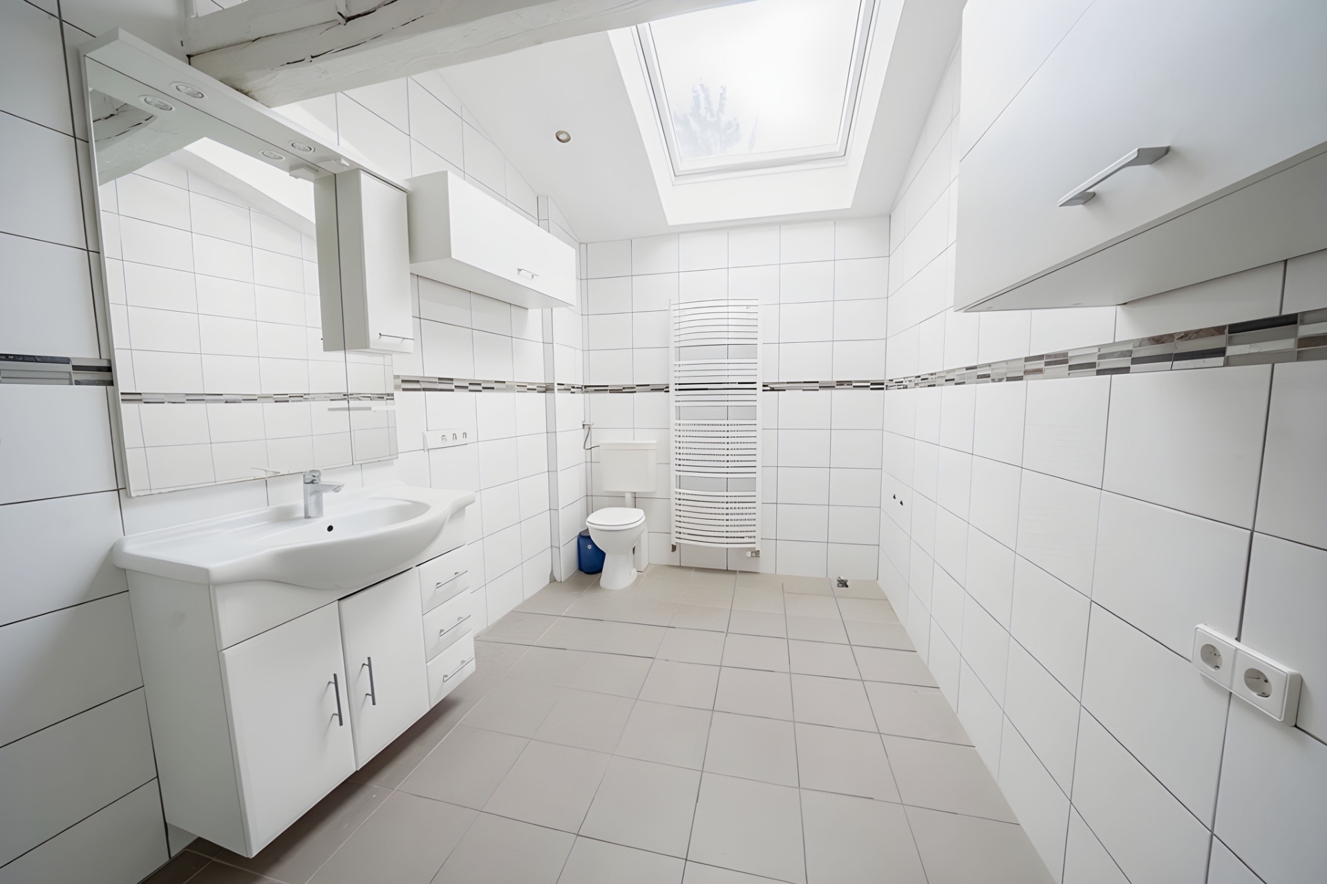 Helles Badezimmer mit weißen Fliesen, Waschbecken, Toilette, Heizkörper und Oberlicht.