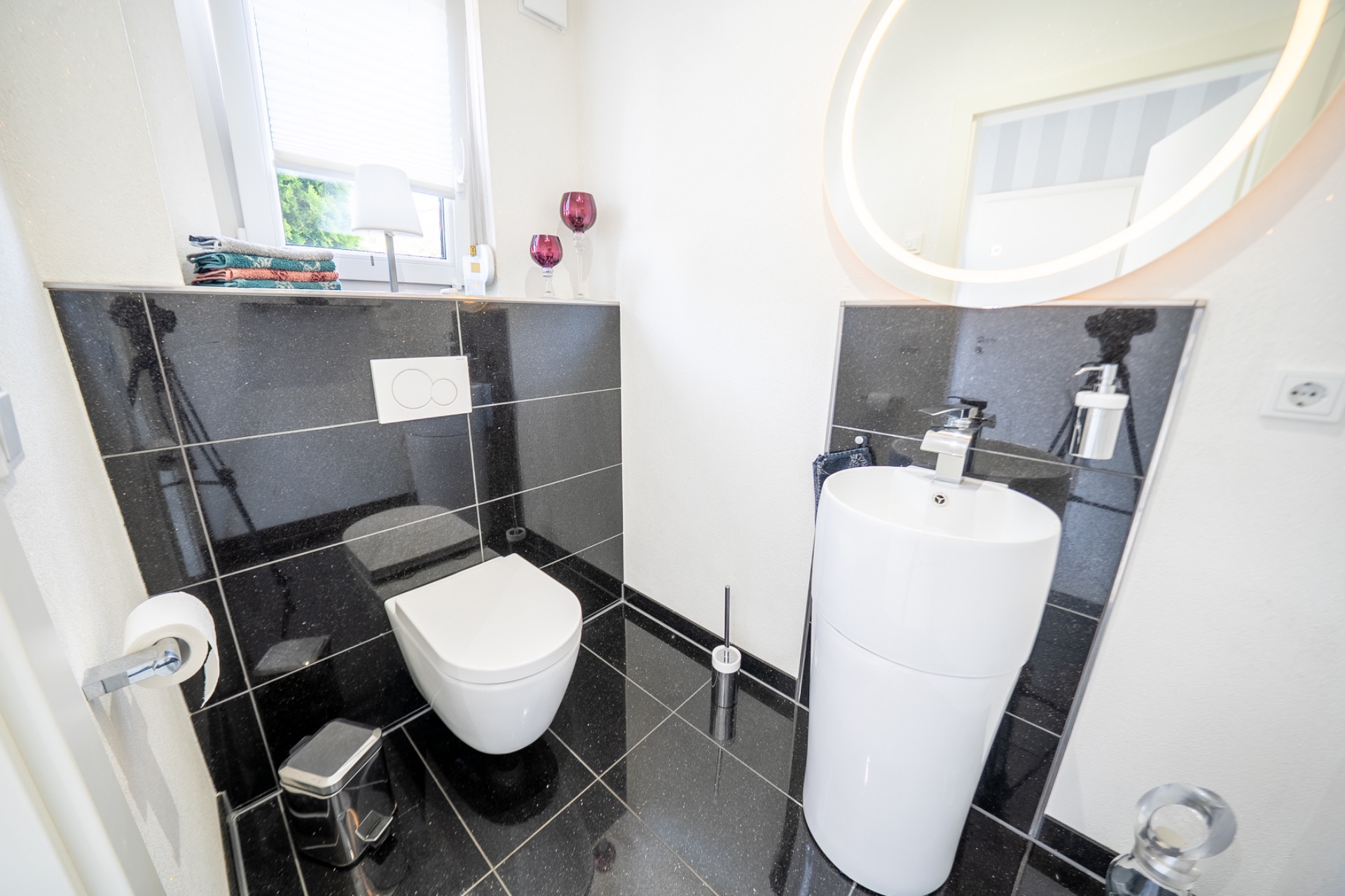 Modernes Badezimmer mit Wandtoilette, Waschbecken, großem Spiegel und schwarzem Fliesenboden.