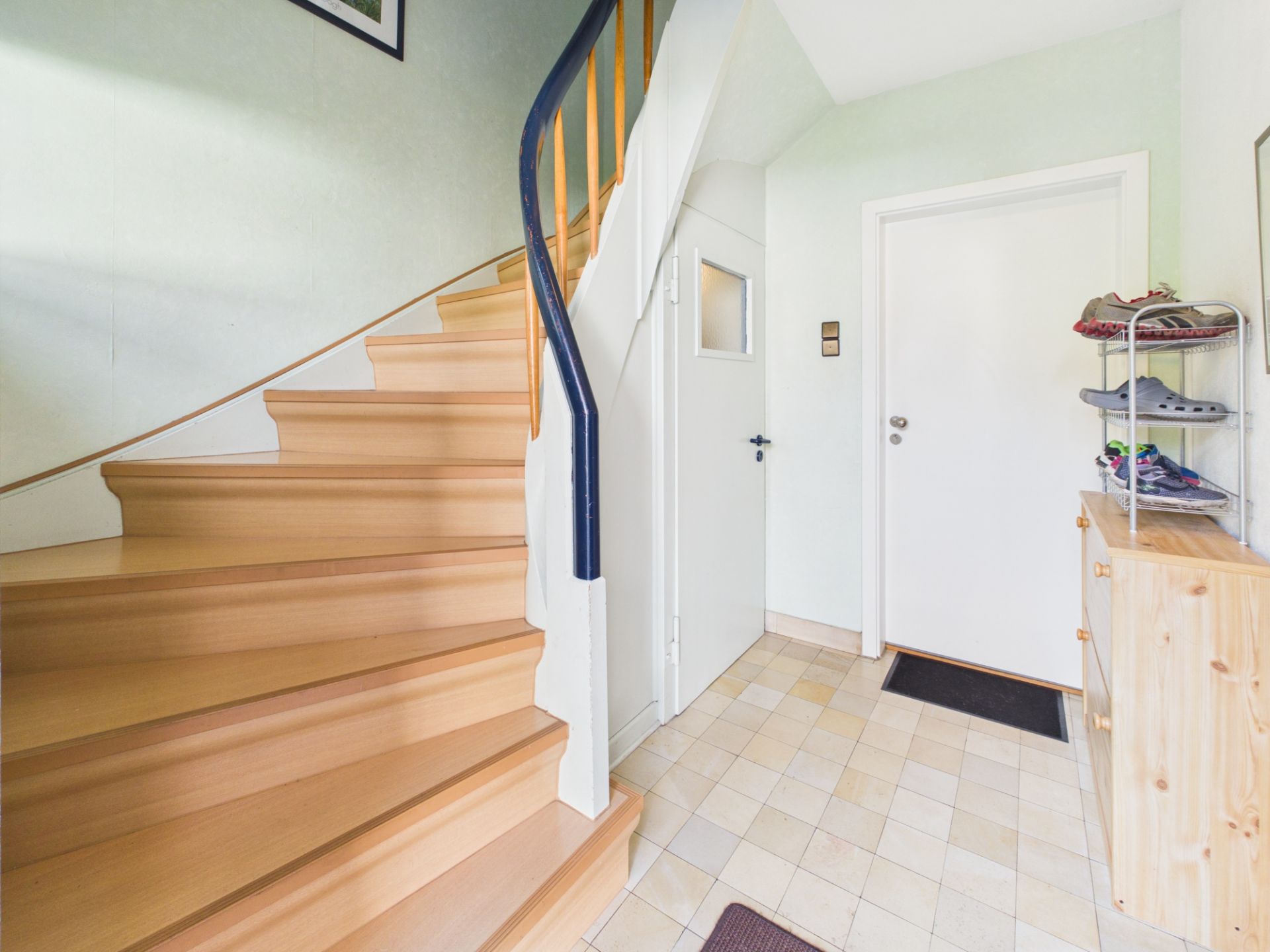 Heller Flur mit Holztreppe, weißer Tür und Schuhregal rechts.