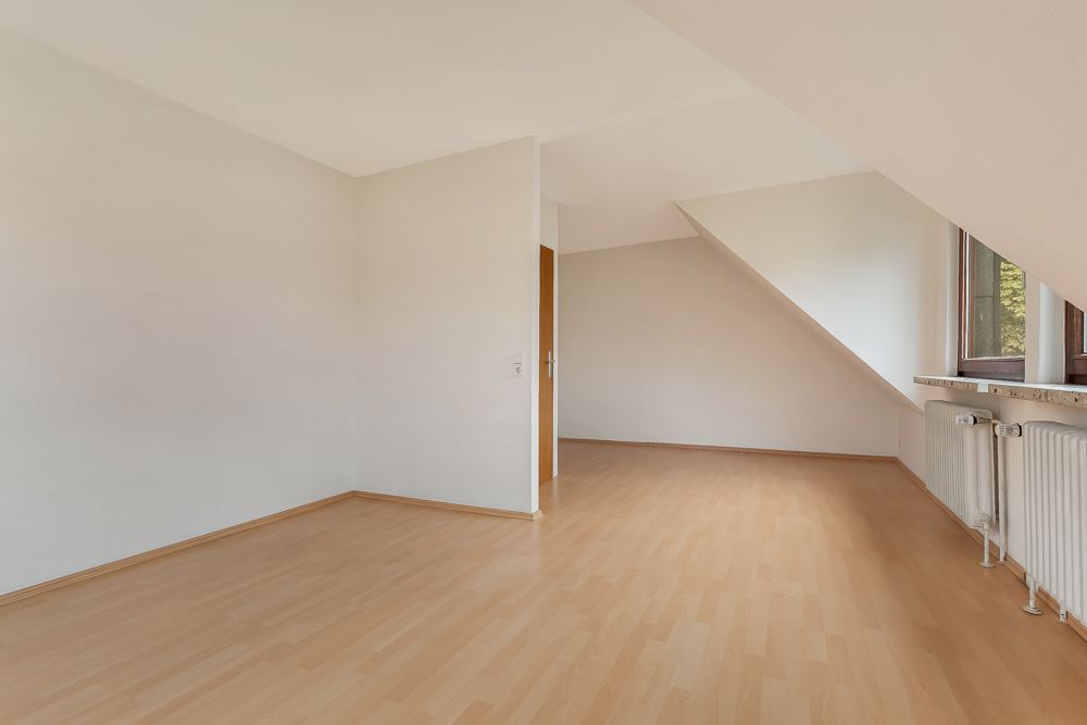 Helles, leeres Zimmer mit schräger Decke, Holzfußboden und Fenstern auf der rechten Seite.