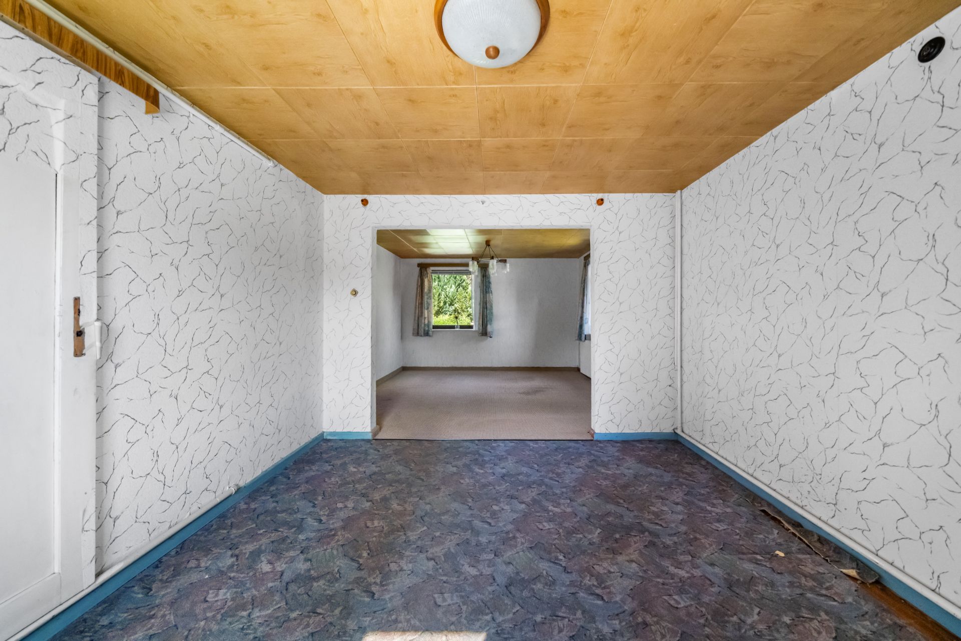 Leerer Wohnraum mit gemusterten Wänden, Holzbalkendecke und Fenster im hinteren Raum.