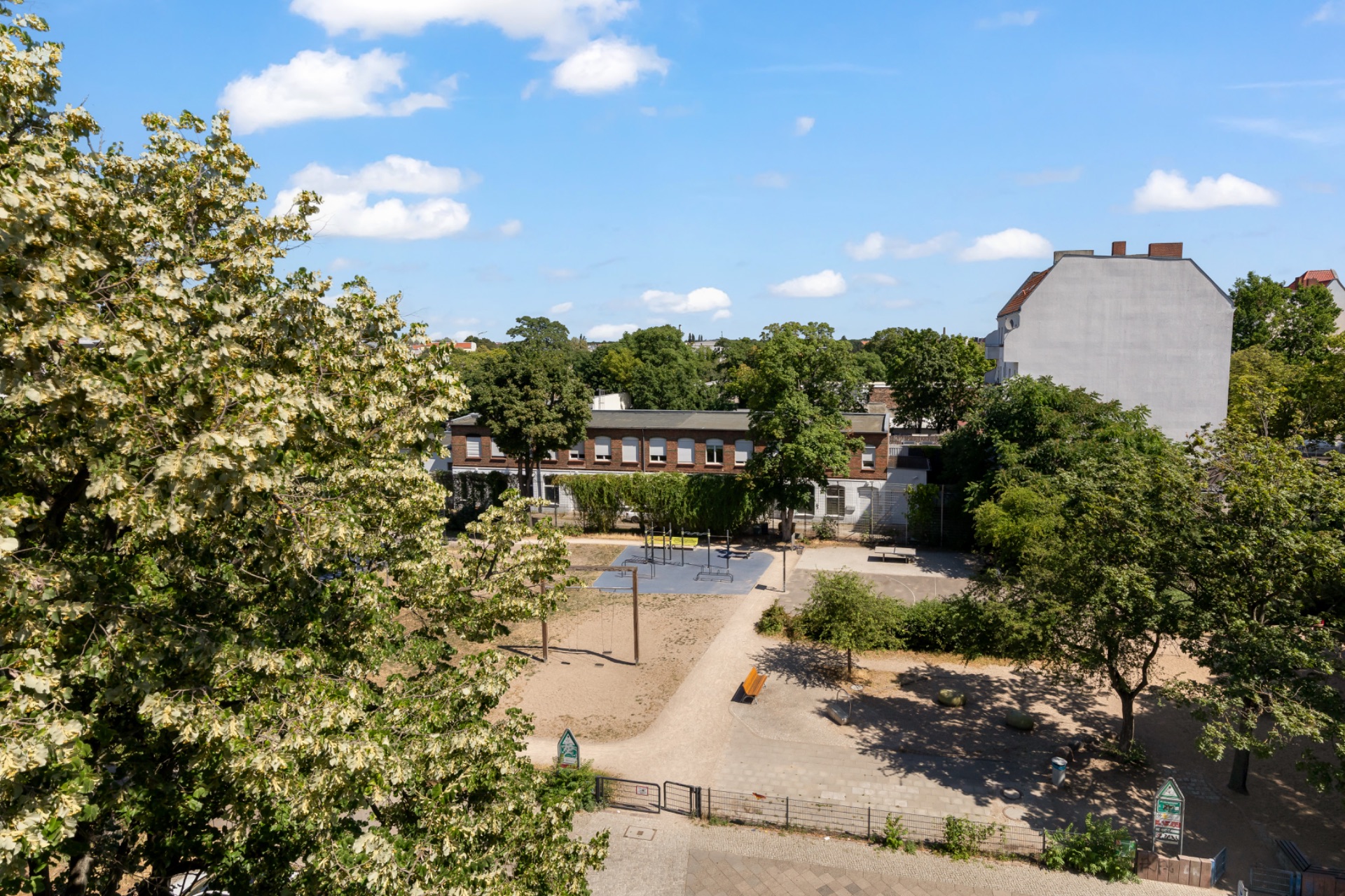 Blick auf einen sonnigen Innenhof mit Bäumen, Bänken und Sandfläche.