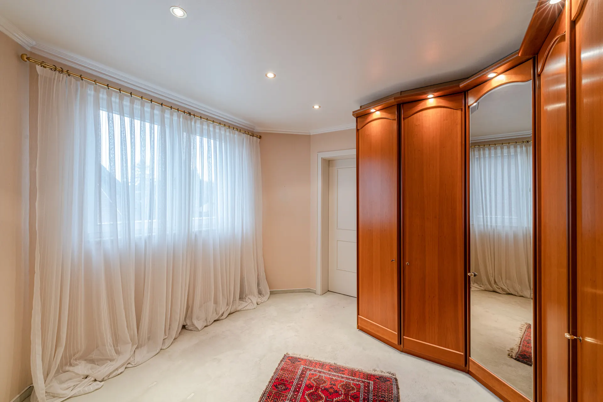 Heller Raum mit großem Fenster, weißen Vorhängen, Holzschrank mit Spiegel und kleinem Teppich.
