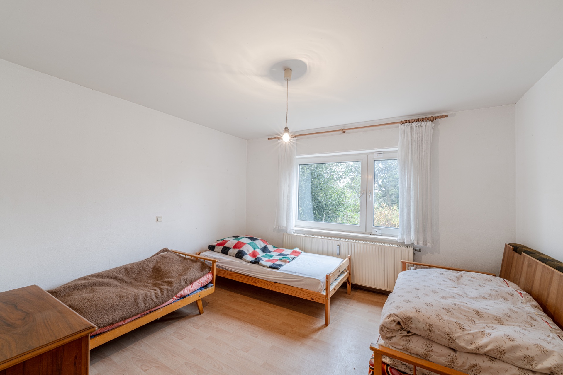 Schlichtes Schlafzimmer mit drei Einzelbetten, Holzboden und großem Fenster mit Vorhang.