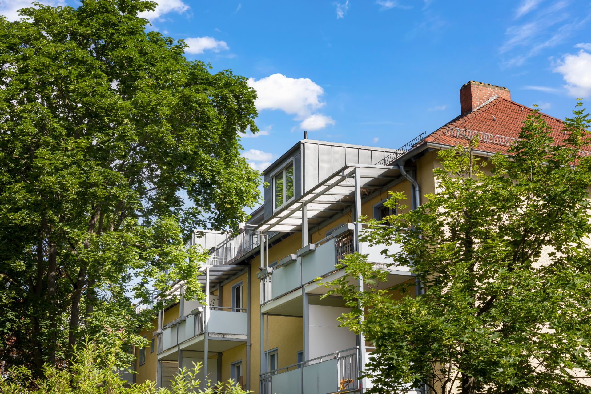Mehrfamilienhaus mit Balkonen, umgeben von grünen Bäumen, unter blauem Himmel.