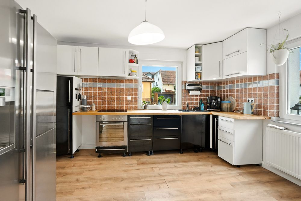 Moderne Küche mit Holzboden, weißen Schränken und braunen Fliesen, große Fenster lassen viel Licht herein.