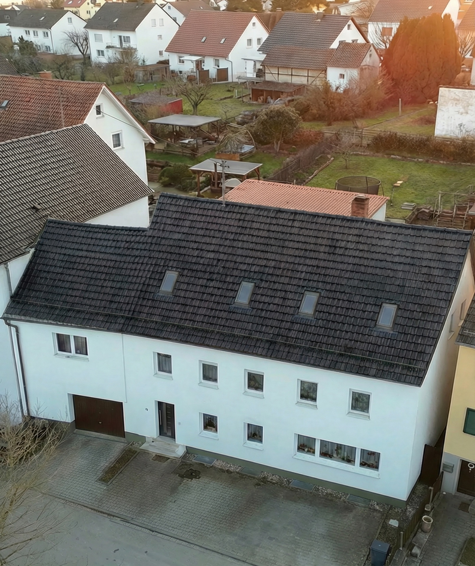 Ein weißes Wohnhaus mit dunklem Satteldach und mehreren Dachfenstern, davor ein gepflasterter Hof.