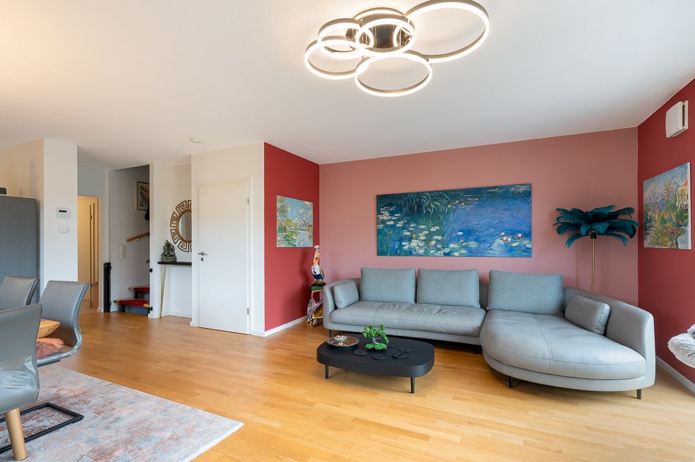 Modernes Wohnzimmer mit grauem Ecksofa, Holzboden und Wandbildern, helle Wände mit rotem Akzent.