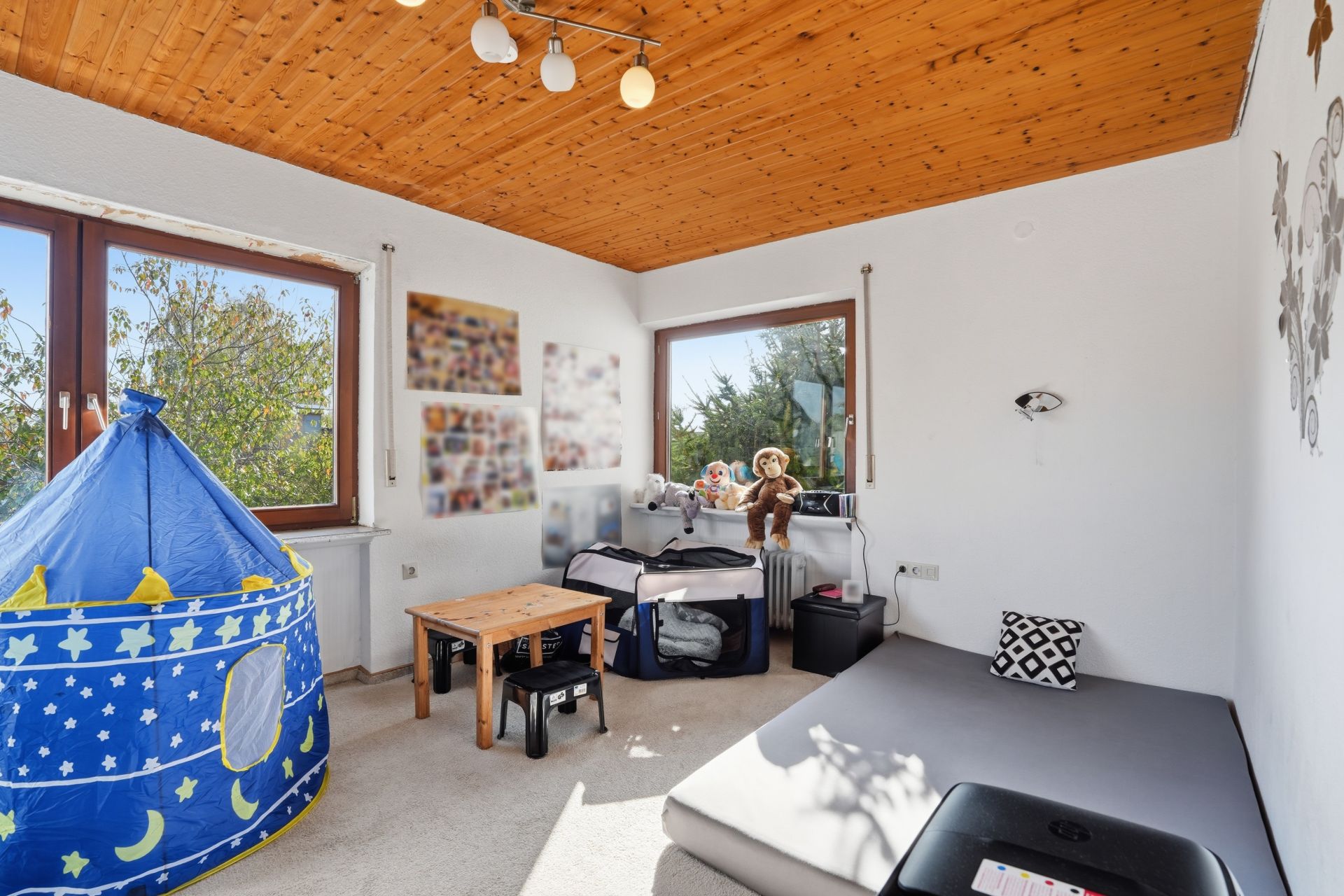 Helles Kinderzimmer mit blauem Spielzelt, Holztisch und großen Fenstern.