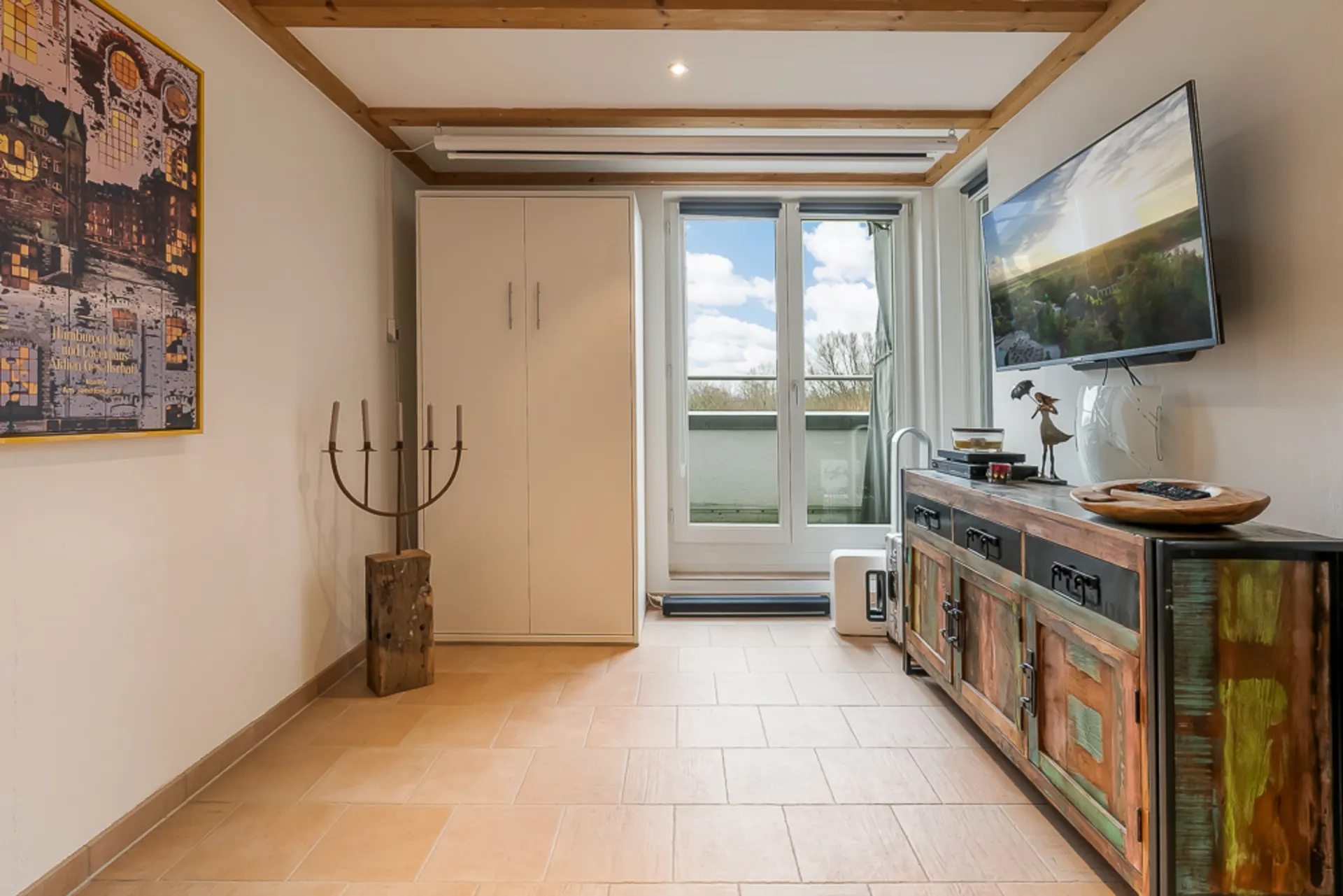 Wohnzimmer mit Fliesenboden, Sideboard, Wandbild, TV und großen Fenstern zur Terrasse.
