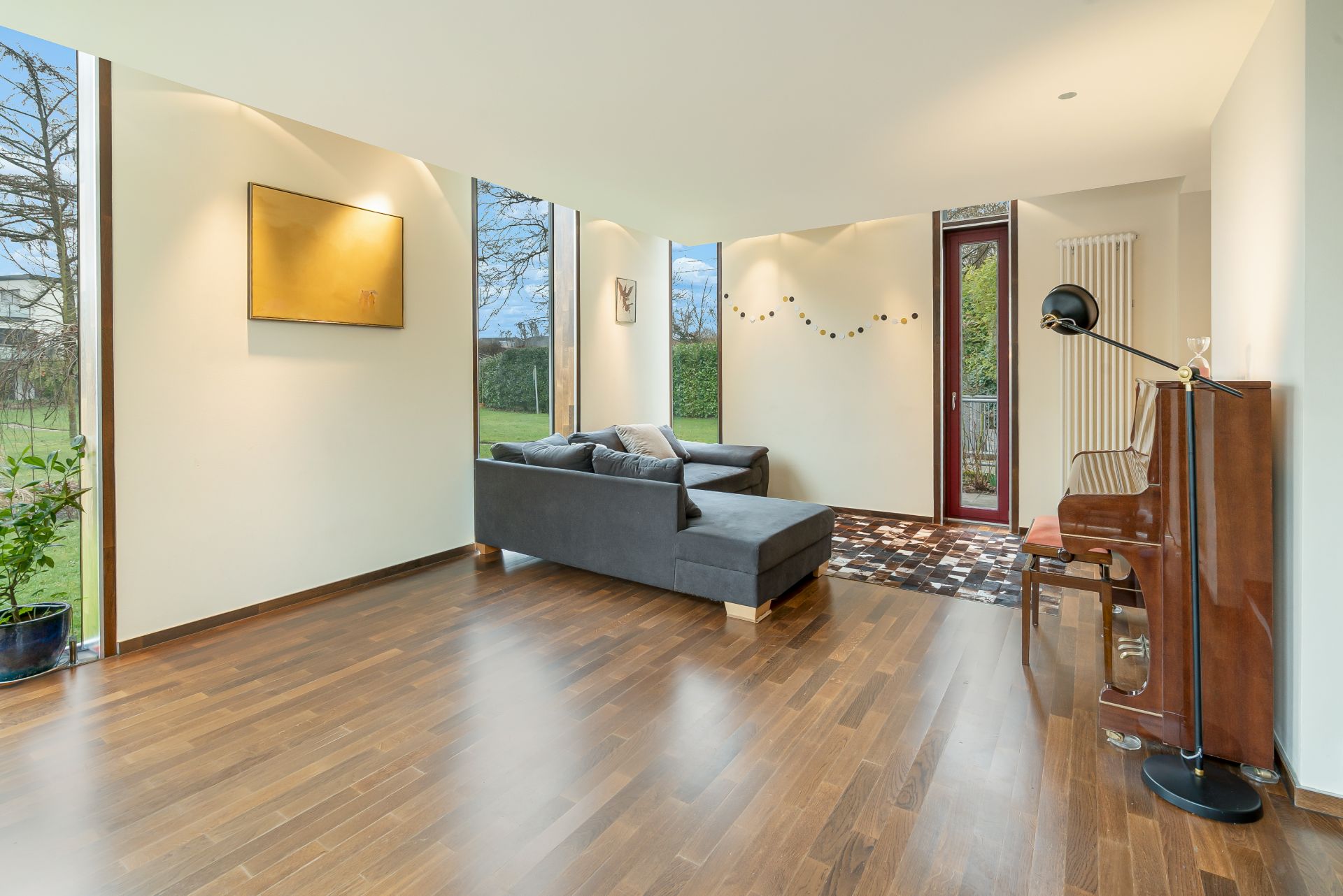 Modernes Wohnzimmer mit Holzboden, Ecksofa, Klavier und großen Fenstern zum Garten.