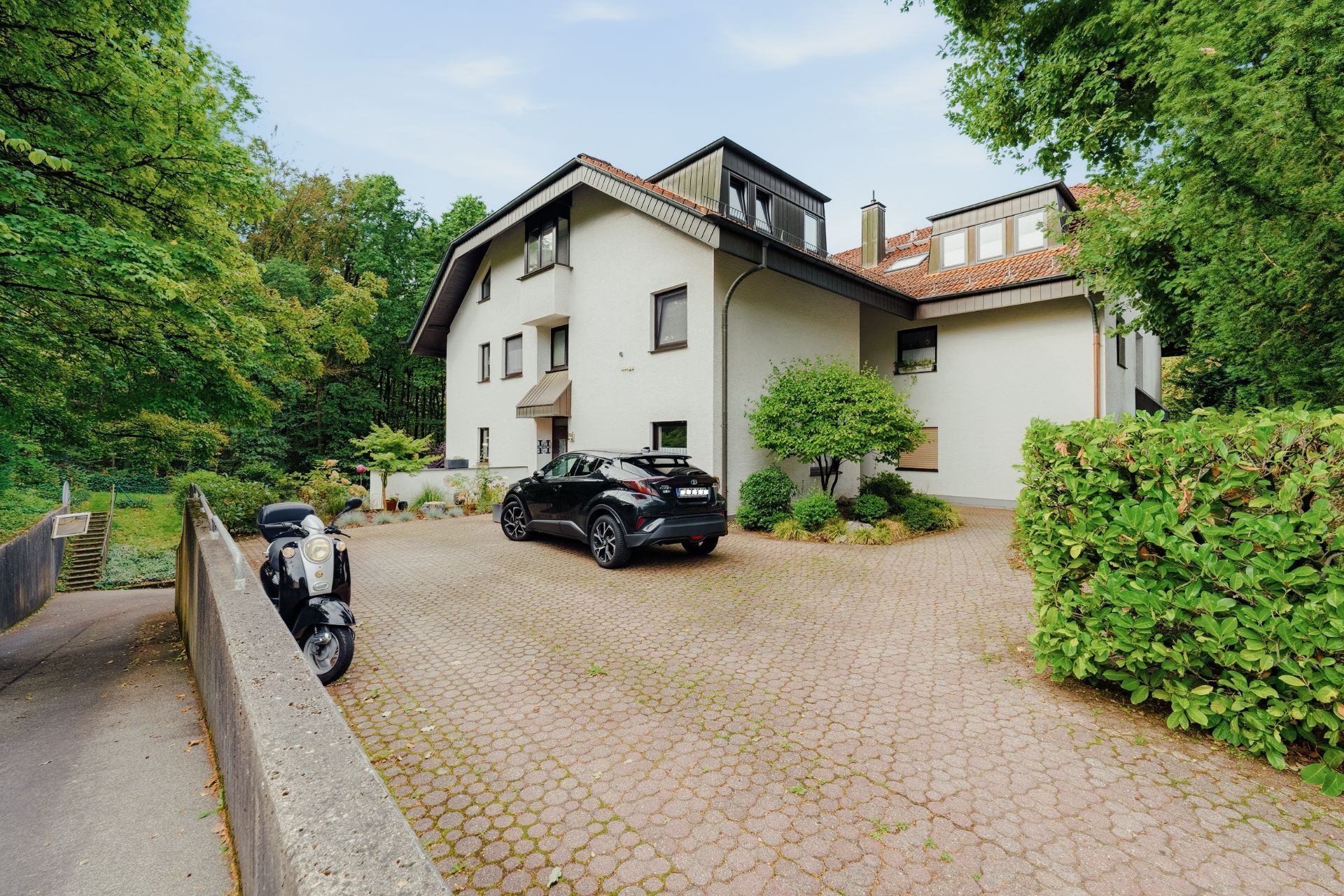 Mehrfamilienhaus mit gepflastertem Hof, Auto und Motorrad im Grünen.