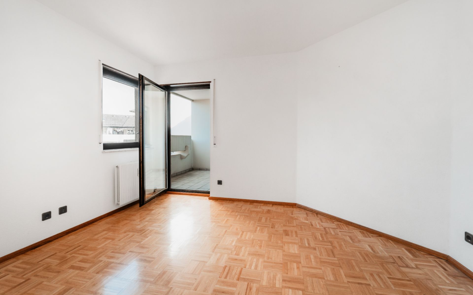 Leeres Zimmer mit Parkettboden, weißem Wänden, Fenster und geöffneter Tür zu einem Balkon.