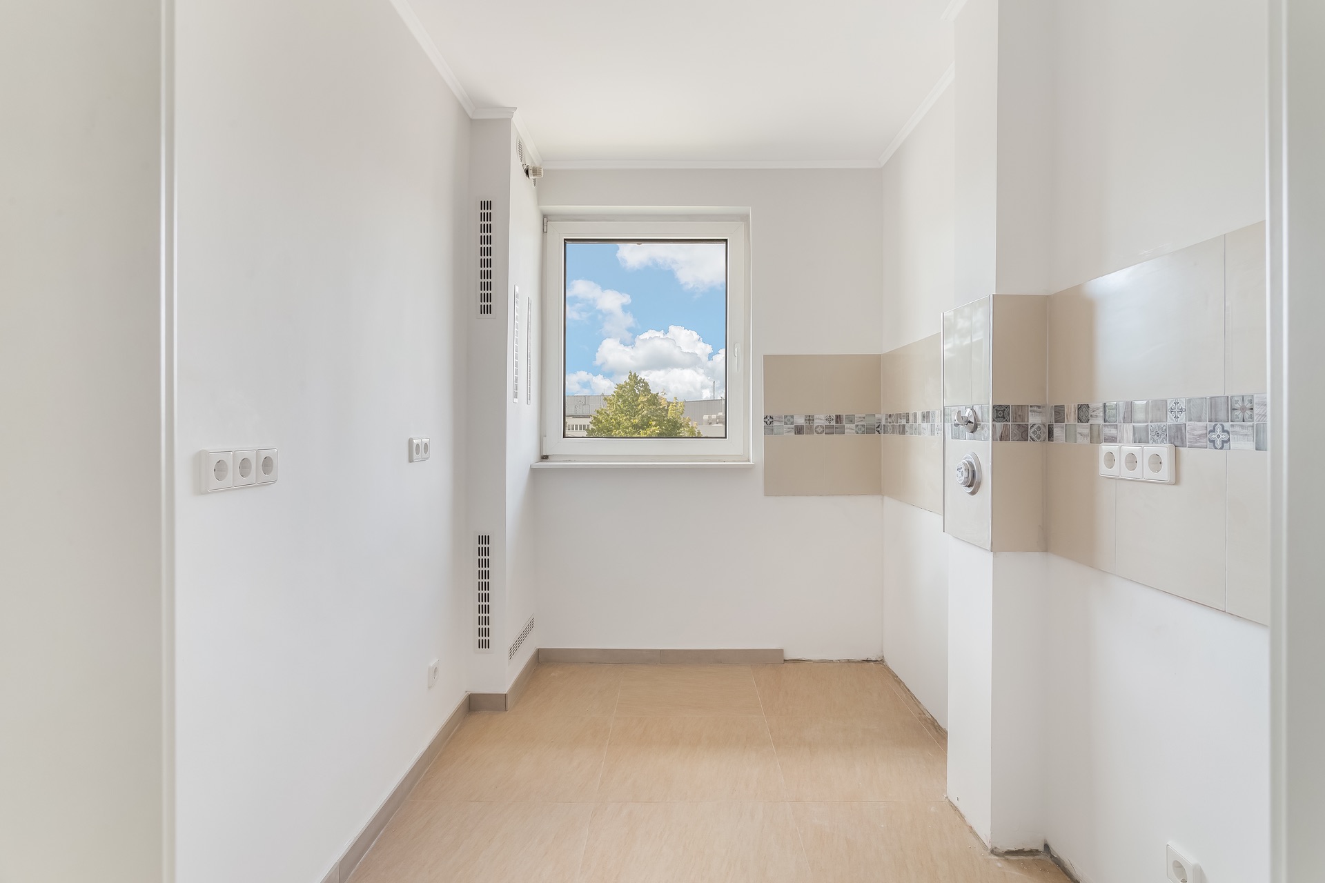 Leerer, heller Raum mit Fliesenboden und Fenster, weiße Wände und Kachelstreifen an einer Wand.