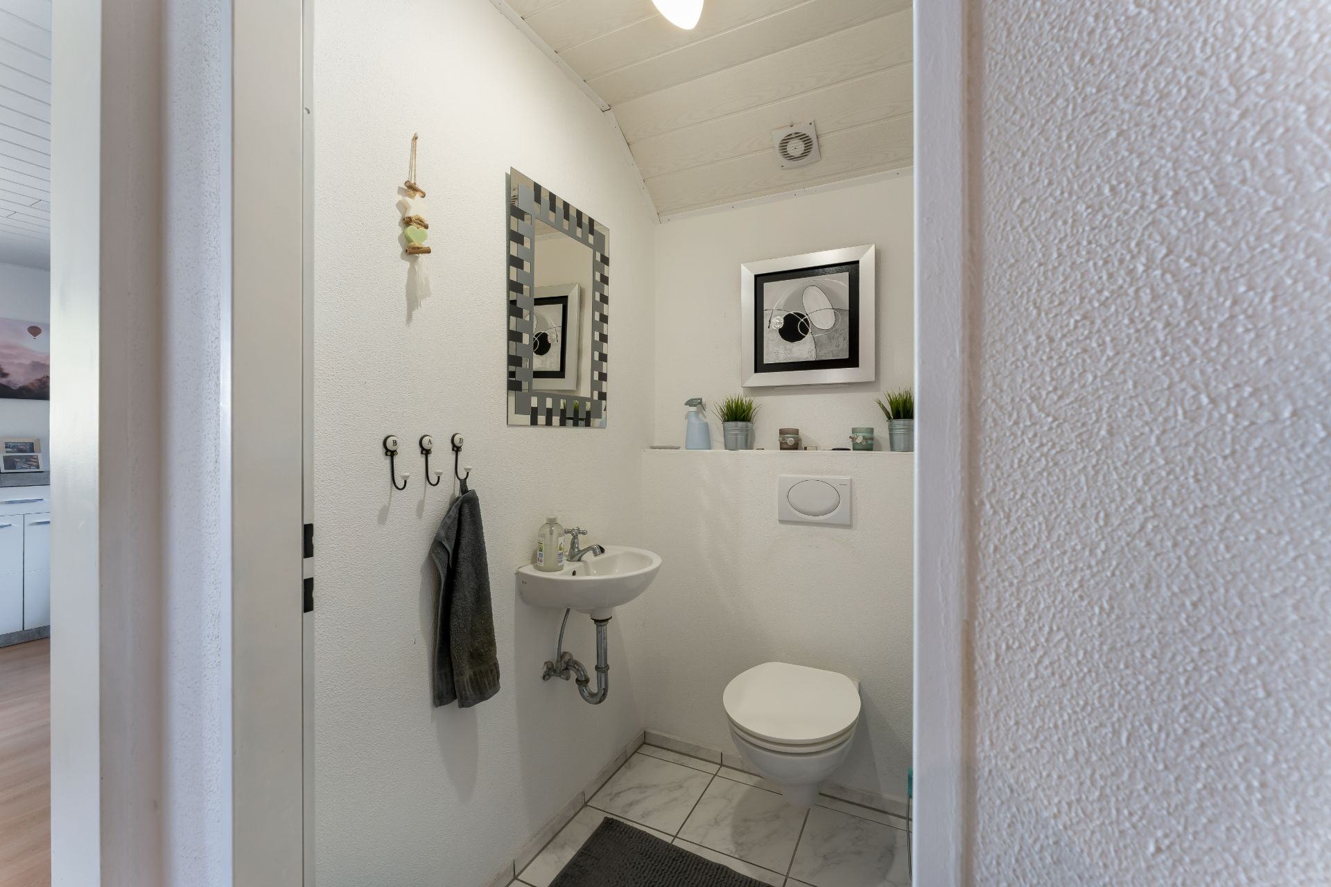 Kleines Badezimmer mit Wandtoilette, Waschbecken, Spiegel und Dekoration auf weißer Ablage.
