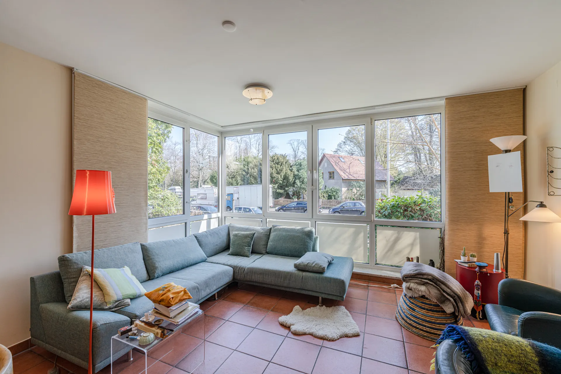 Helles Wohnzimmer mit Ecksofa, großen Fenstern, roten Fliesen und mehreren Stehlampen.