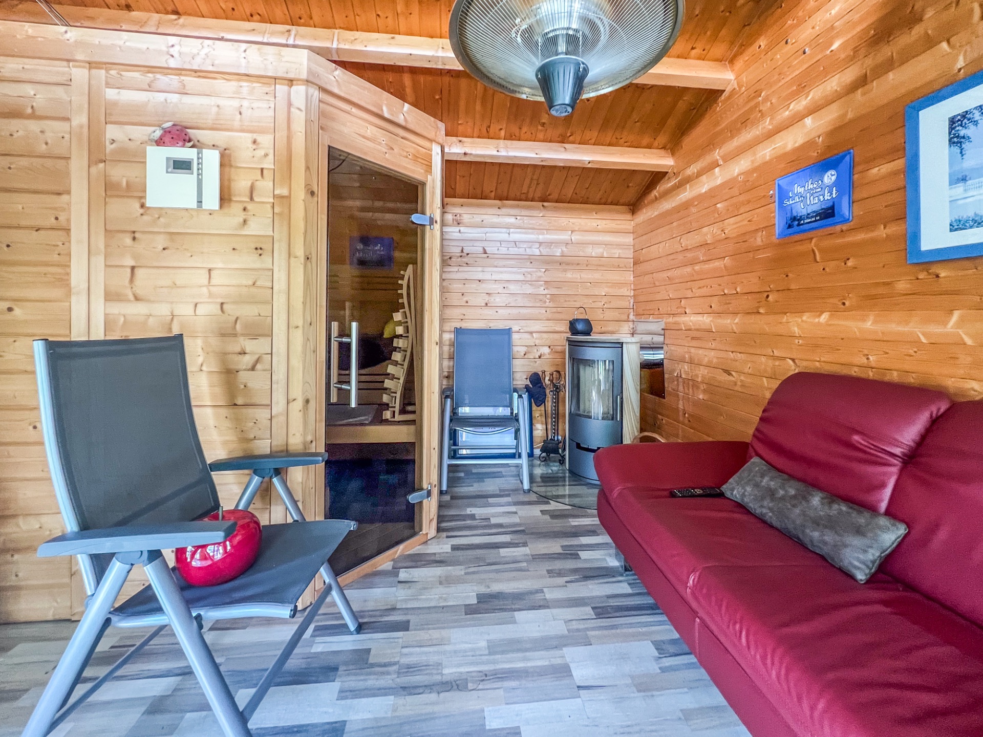 Wohnraum mit Holzverkleidung, rotem Sofa, Saunakabine und Kaminofen.