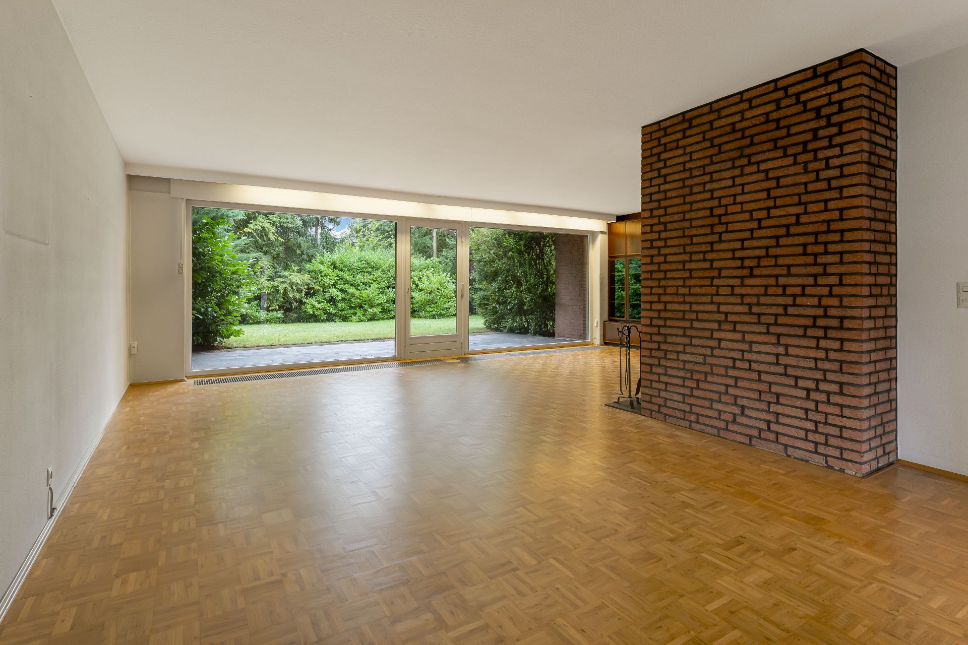 Großes, leeres Wohnzimmer mit Parkettboden, Backsteinwand und bodentiefen Fenstern zum Garten.