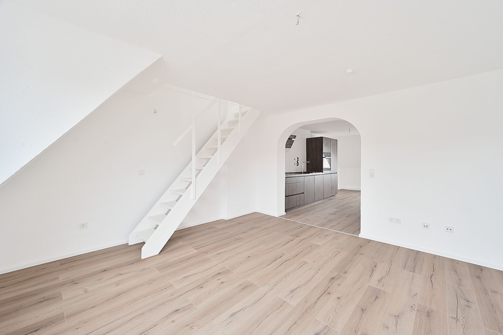 Helles Zimmer mit Holzfußboden, weißer Treppe und Durchgang zur modernen Küche.