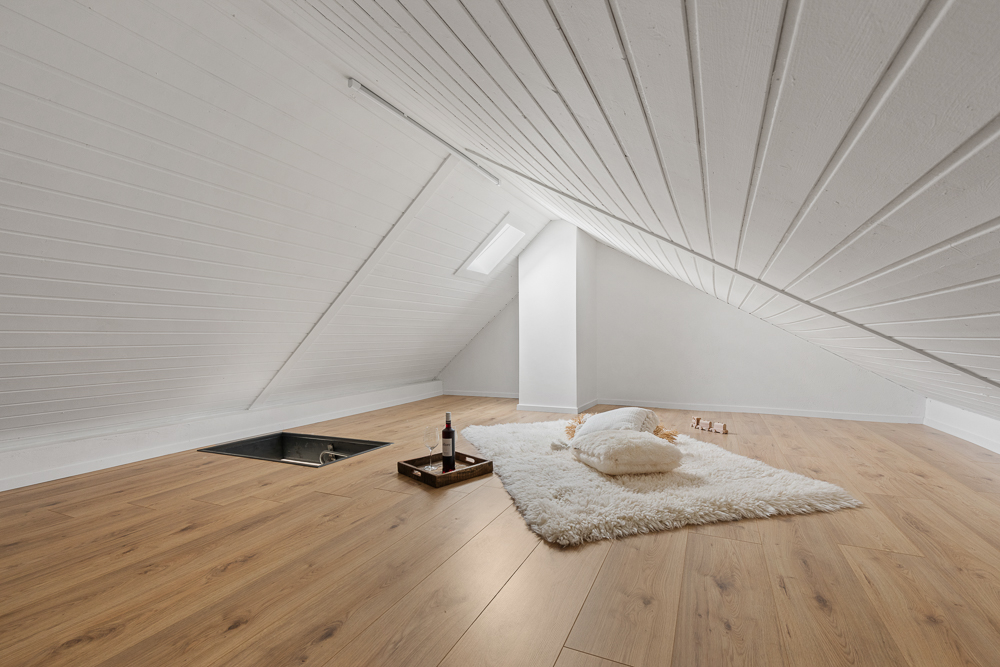Heller Dachbodenraum mit schräger Decke, Holzfußboden, Teppich, Kissen und einem Tablett mit Weinflasche.