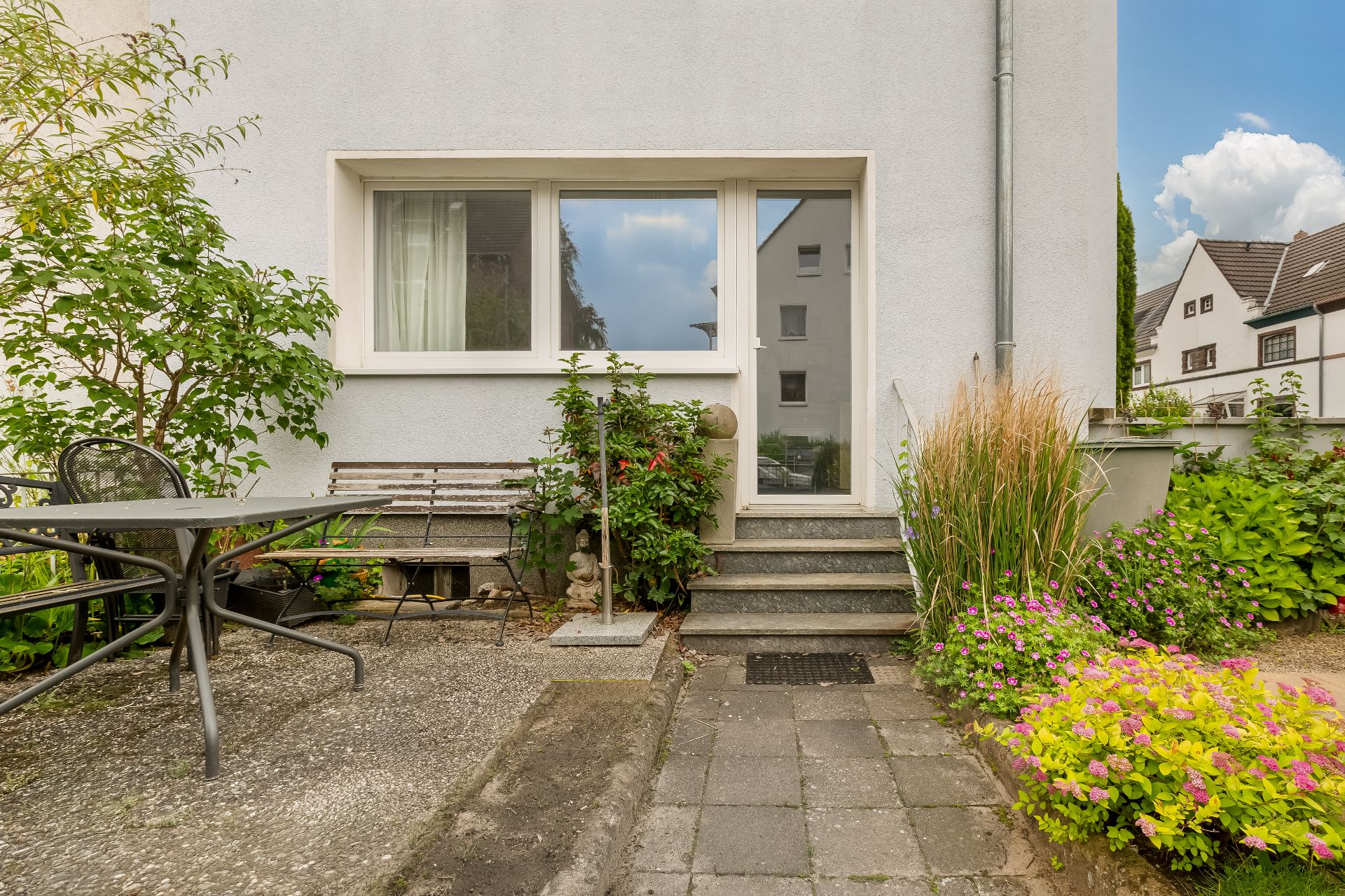 Hauseingang mit Glas­tür, Treppe, Sitzbank, Tisch, Stuhl und blühenden Pflanzen im Garten.
