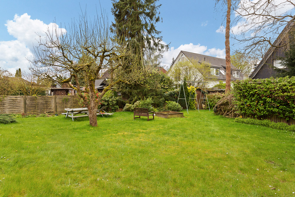 Grüner Garten mit Bäumen, Picknicktisch, Bank, Schaukel und Beet, umgeben von Hecken und Häusern.