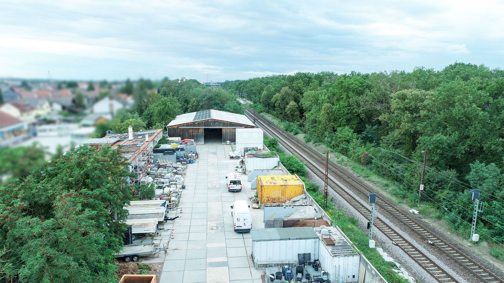 Lagerplatz mit Containern und Materialien neben Bahngleisen und Bäumen.