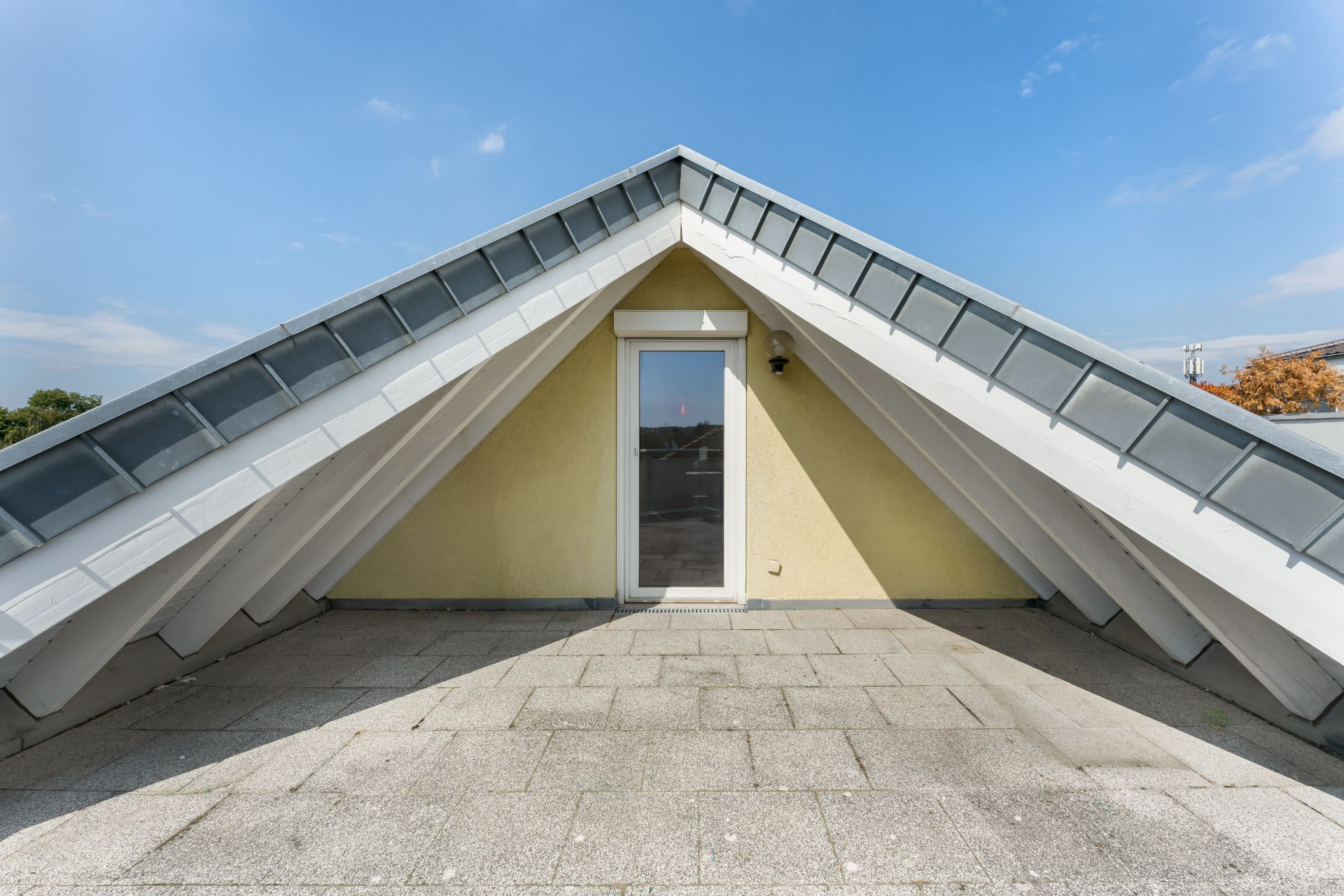 Dachterrasse mit Schrägdach, Glastür und gefliestem Boden, Himmel im Hintergrund.