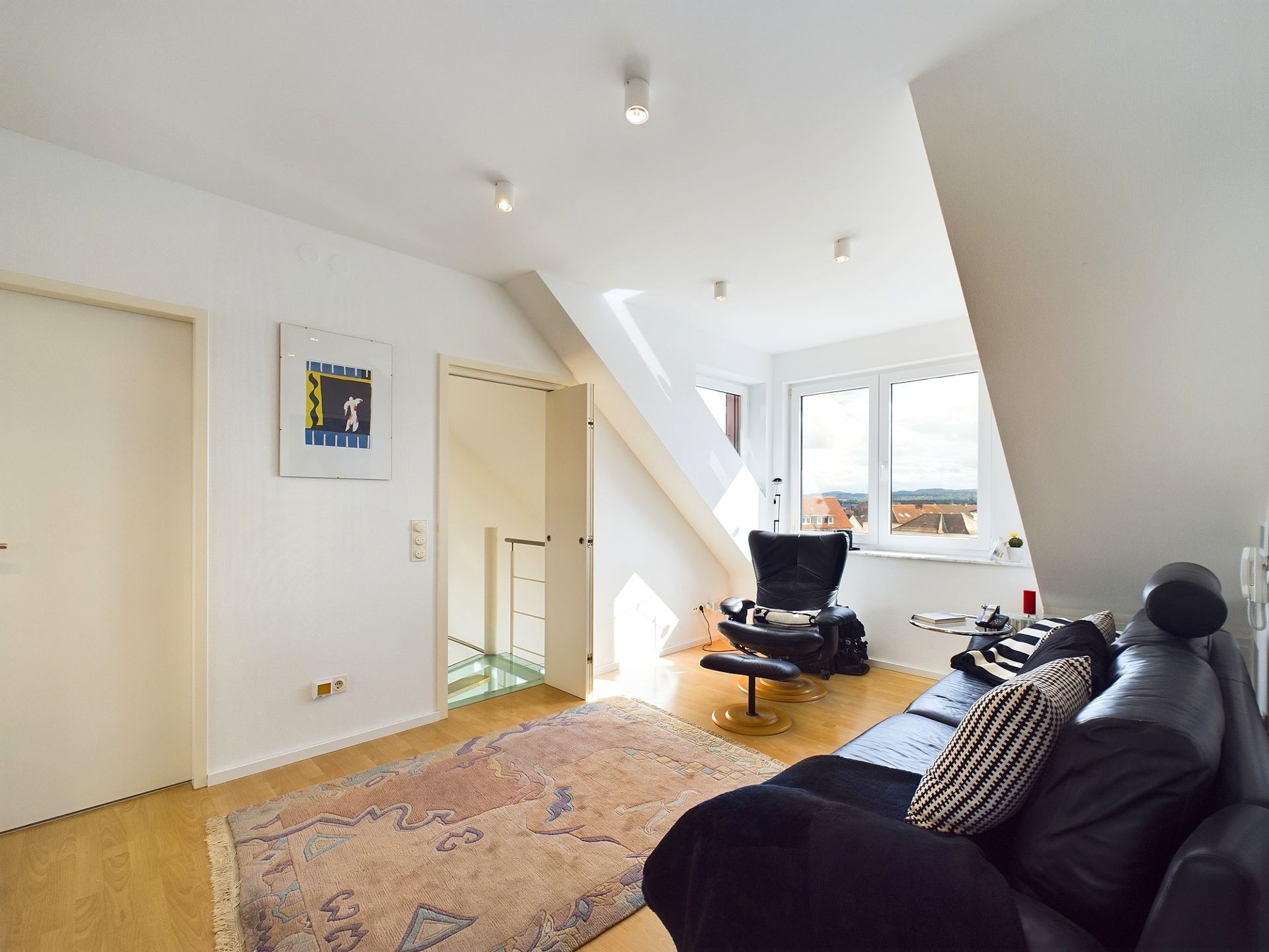 Helles Wohnzimmer mit schwarzem Sofa, Sessel, Teppich und Dachfenster.
