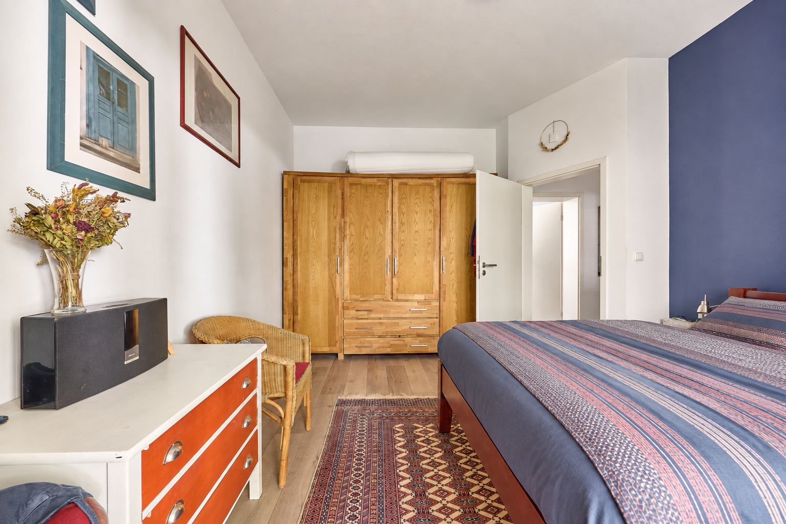 Schlafzimmer mit Doppelbett, Holzschrank, Teppich, Stuhl und Kommode mit Vase.