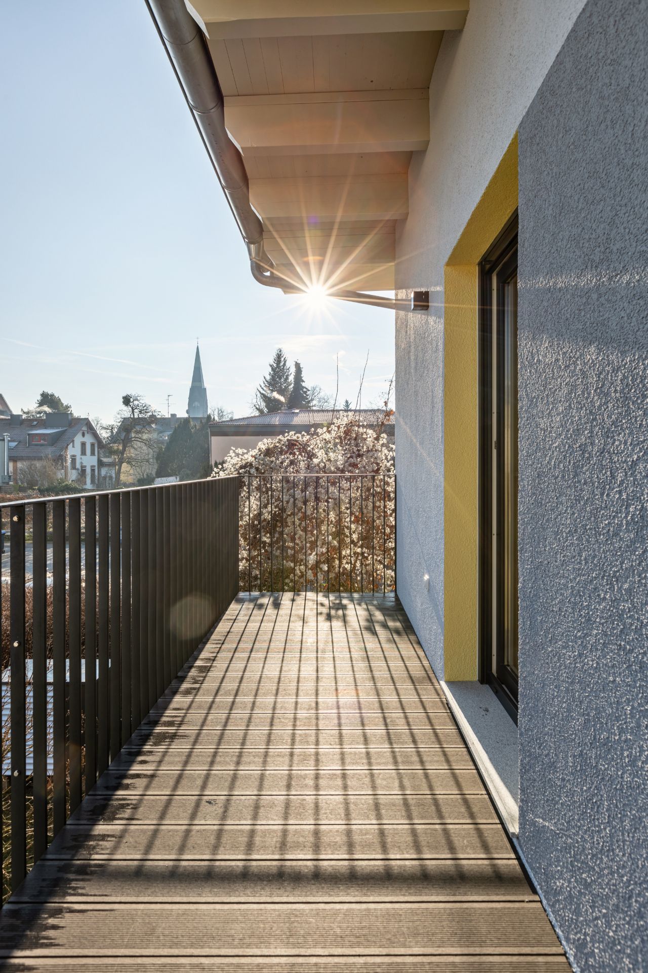 Balkon mit Holzboden und Metallgeländer an einem Wohnhaus, Sonnenstrahlen im Hintergrund.