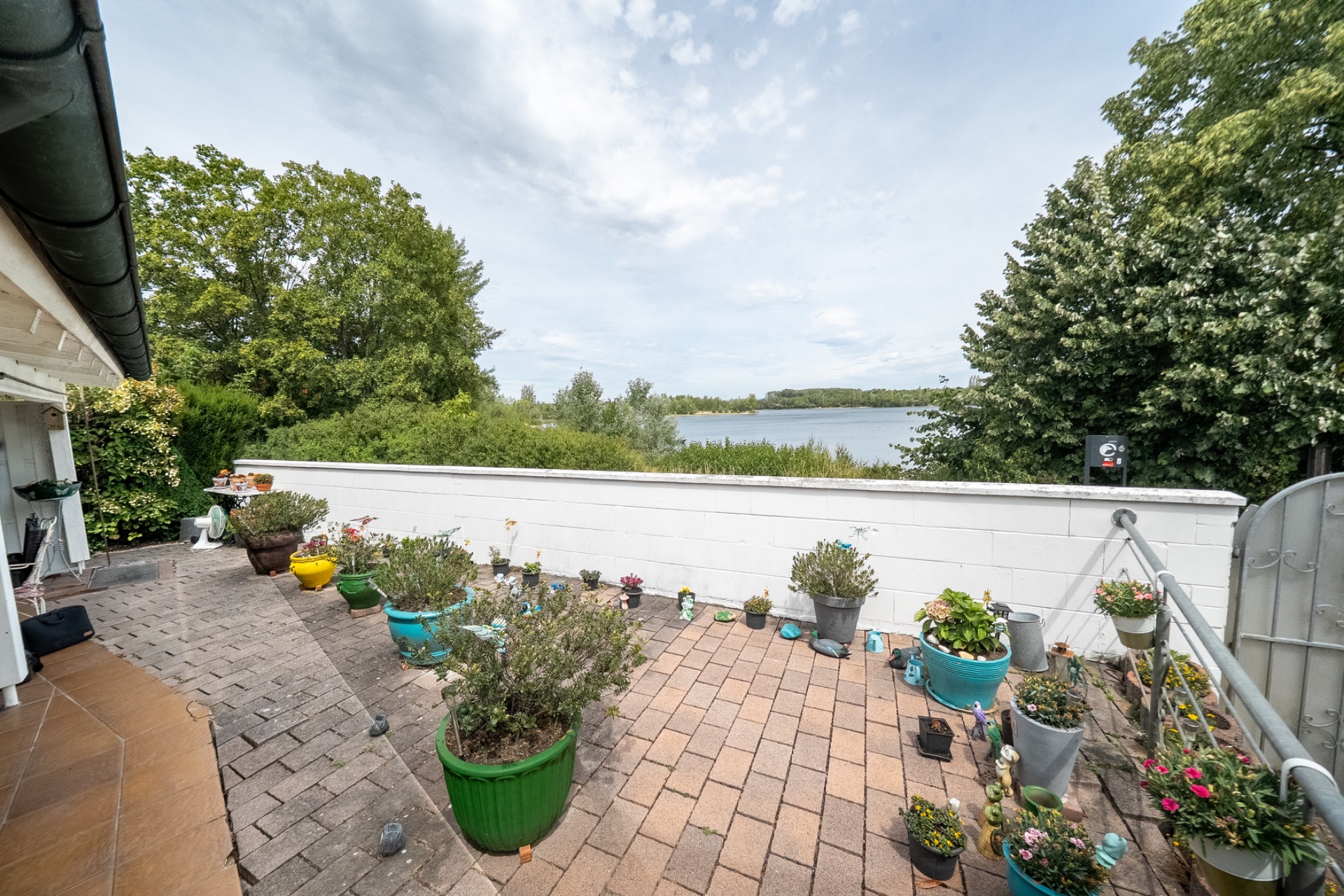 Terrasse mit vielen Blumentöpfen, Blick auf See und Bäume im Hintergrund.