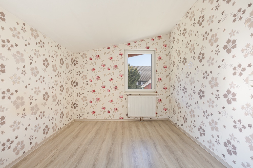 Leeres Zimmer mit heller Blumen-Tapete, Fenster und Heizkörper, Boden aus hellem Holz.
