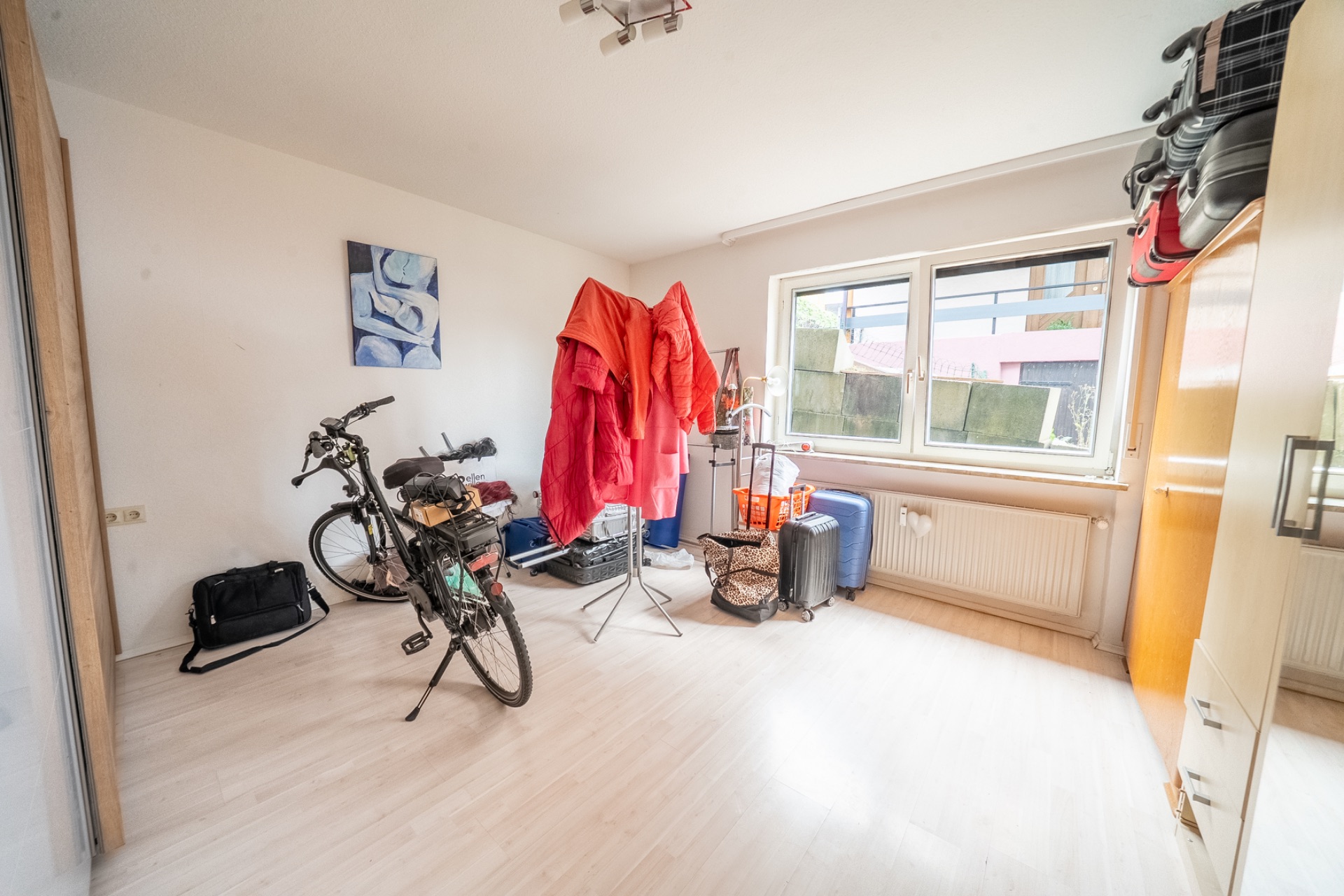 Helles Zimmer mit Fahrrad, Kleiderständer, Koffern und hellen Möbeln vor einem Fenster.