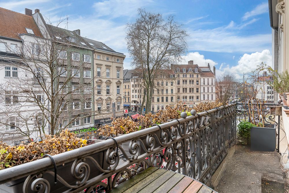 Balkon mit Metallgeländer, Blick auf Straße und mehrstöckige Wohnhäuser, einige Pflanzen auf dem Balkon.