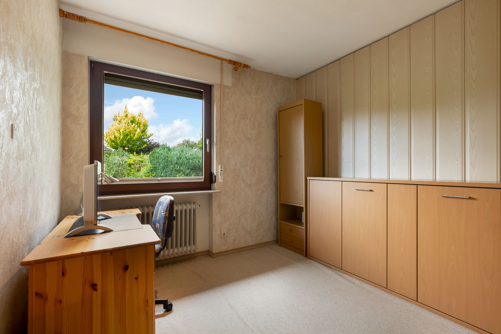 Helles Zimmer mit Holzschreibtisch, Stuhl, Holzschränken und großem Fenster mit Blick ins Grüne.