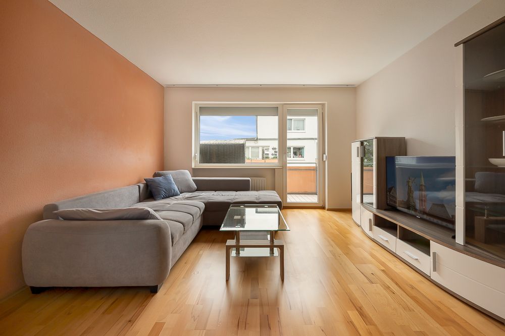 Wohnzimmer mit grauem Ecksofa, Glastisch, Fernseher und Holzfußboden.