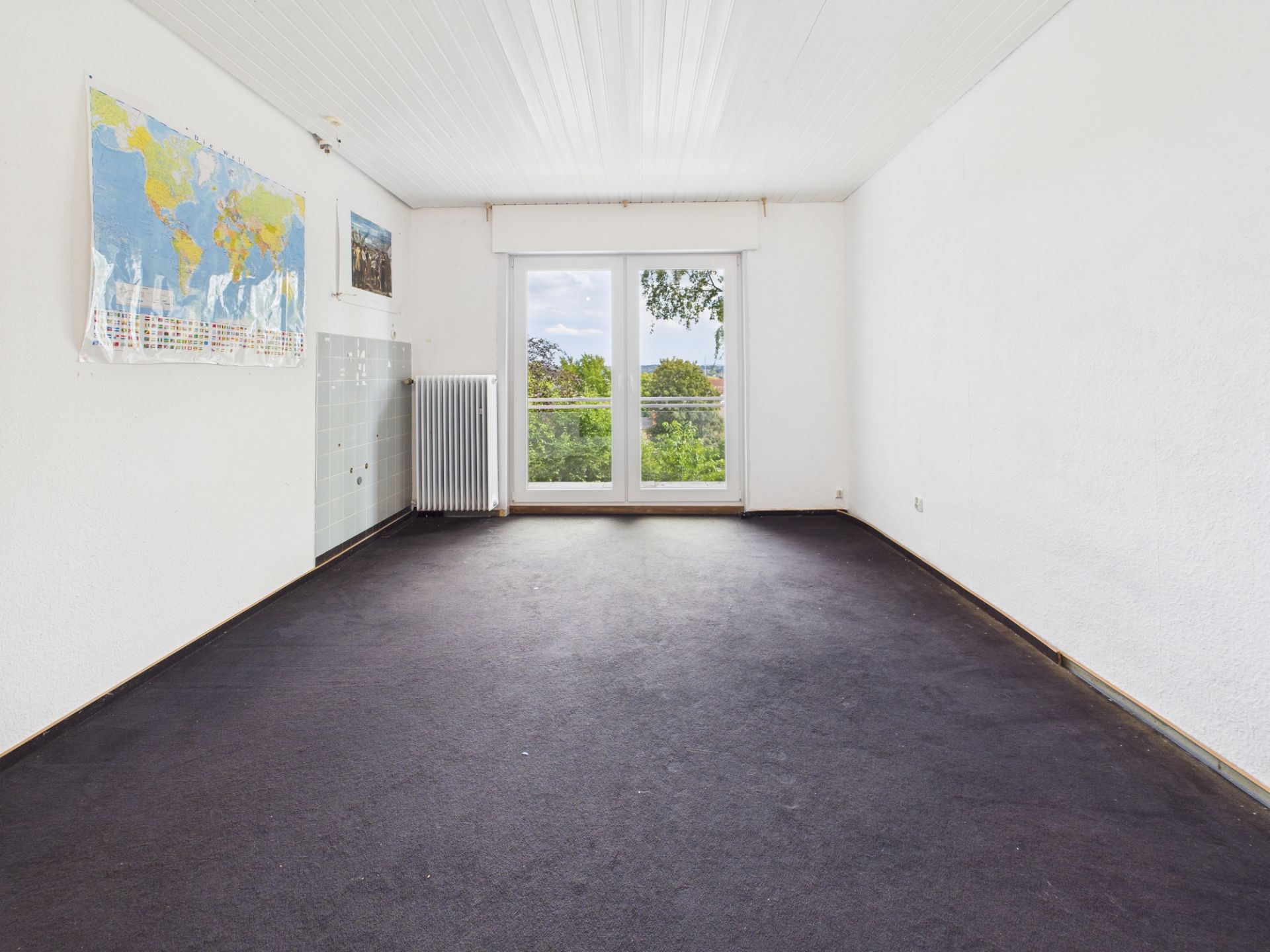 Leeres Zimmer mit dunklem Teppich, Weltkarte an der Wand und großer Fensterfront mit Blick ins Grüne.