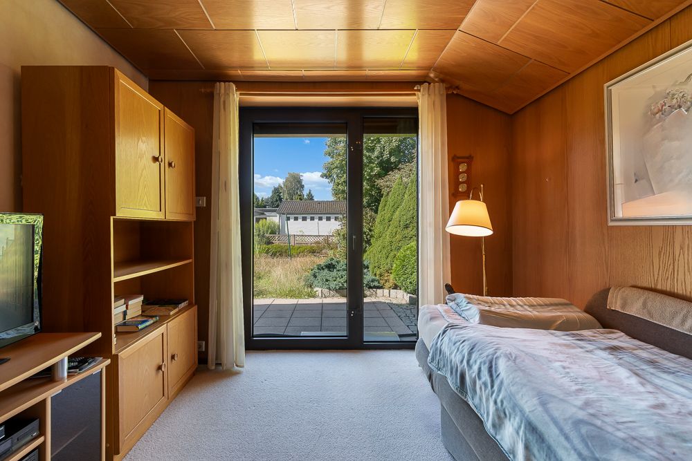 Wohnzimmer mit Einzelbett, Holzschrank, Flügeltür und Blick auf Terrasse und Garten.