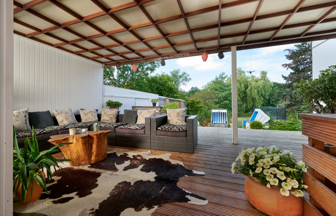 Überdachte Terrasse mit Sitzgruppe, Holztisch, Teppich, Pflanzen und Blick auf einen Garten.