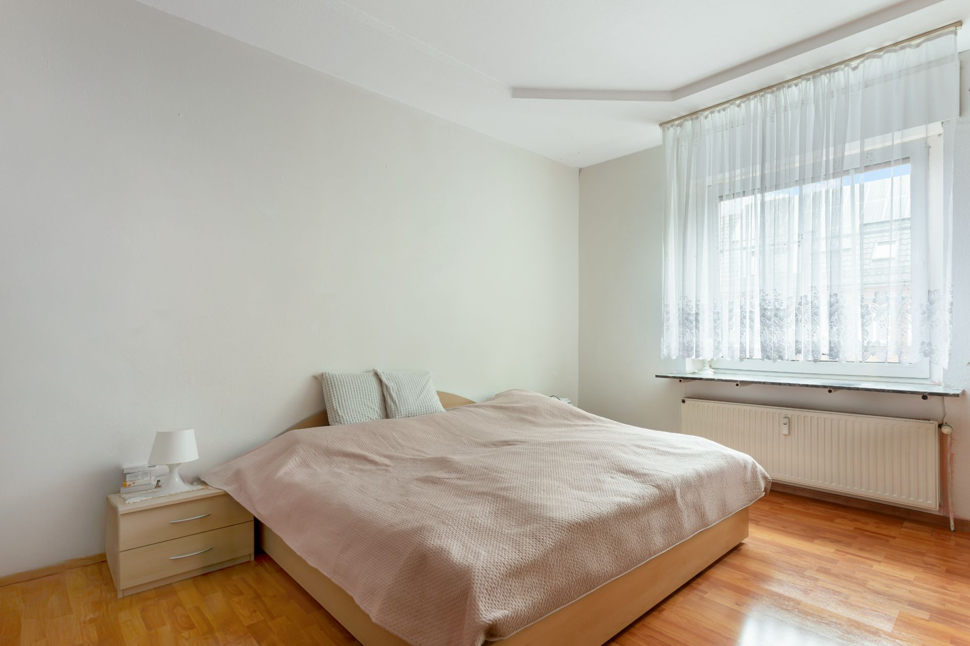 Helles Schlafzimmer mit Doppelbett, Nachttisch, Lampe und großem Fenster mit weißen Vorhängen.