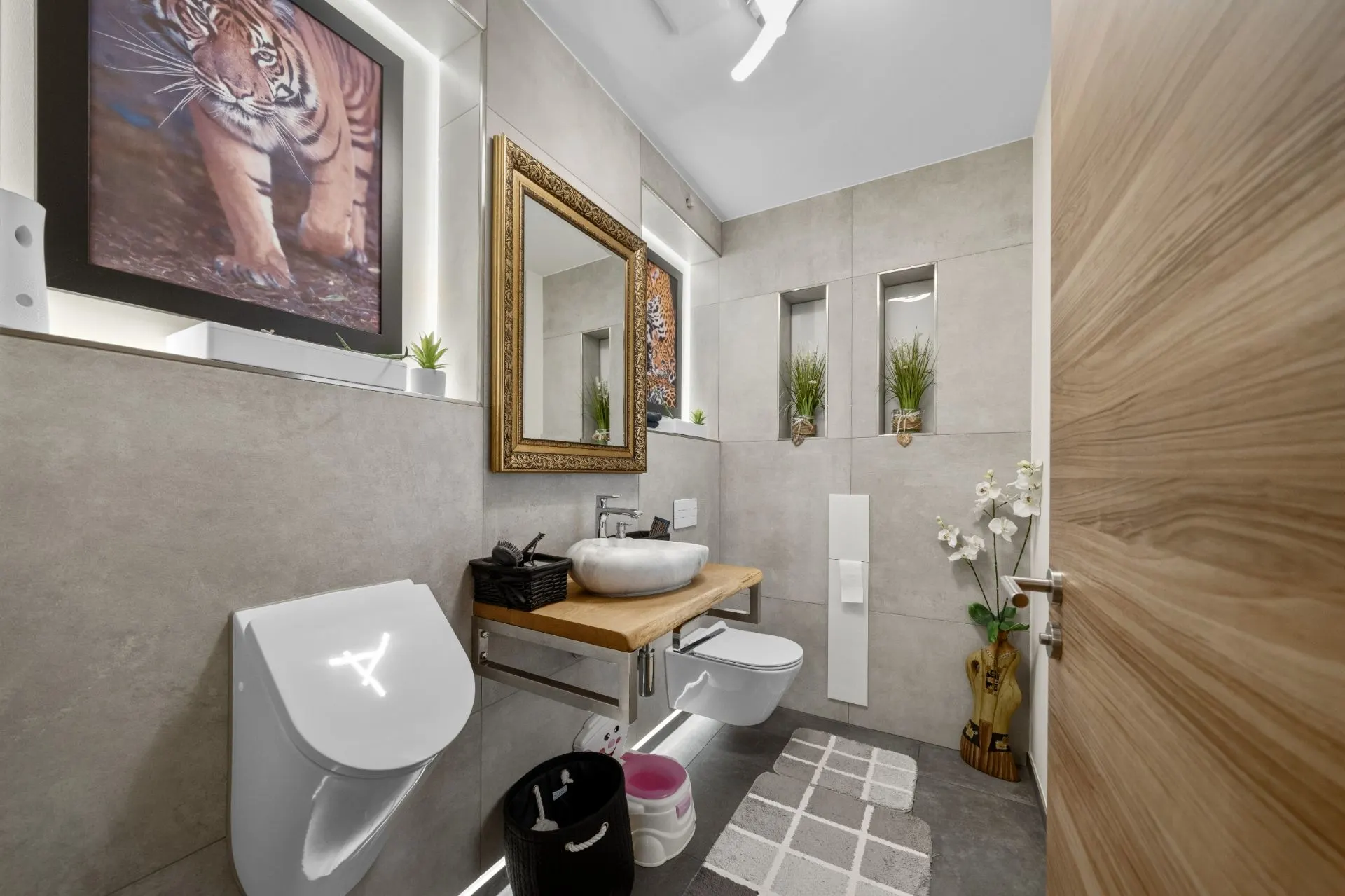 Modernes Badezimmer mit Waschbecken, Spiegel, WC, Urinal, Pflanzendekoration und Tigerbild an der Wand.