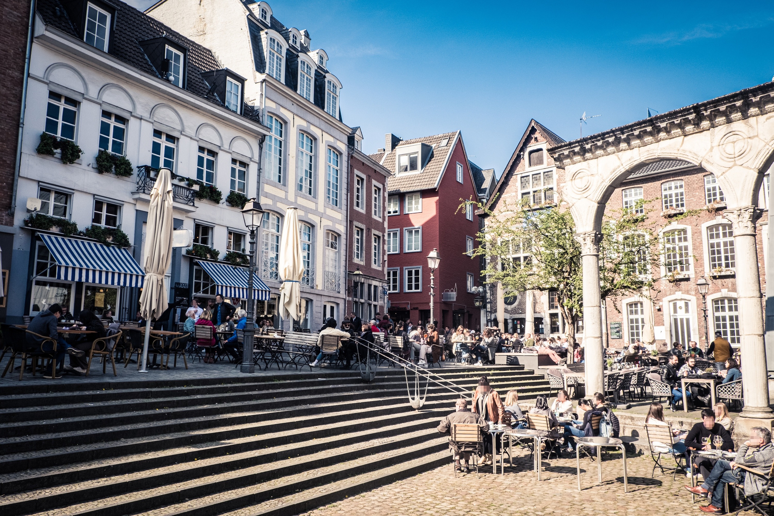Menschen sitzen auf einer Terrasse mit Stufen vor historischen Wohnhäusern.