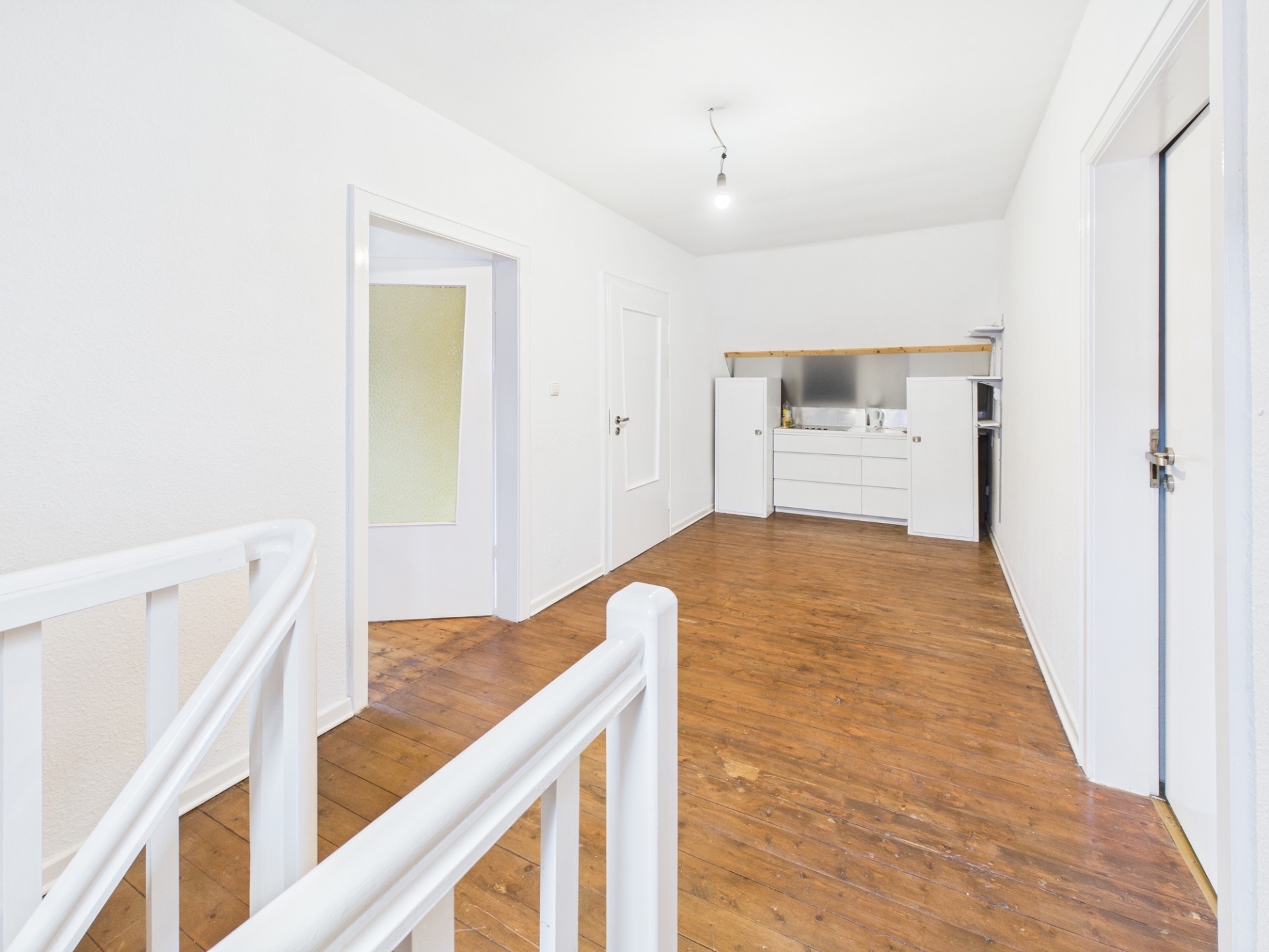 Heller Raum mit Holzfußboden, weißer Treppe und kleiner Küchenzeile im Hintergrund.