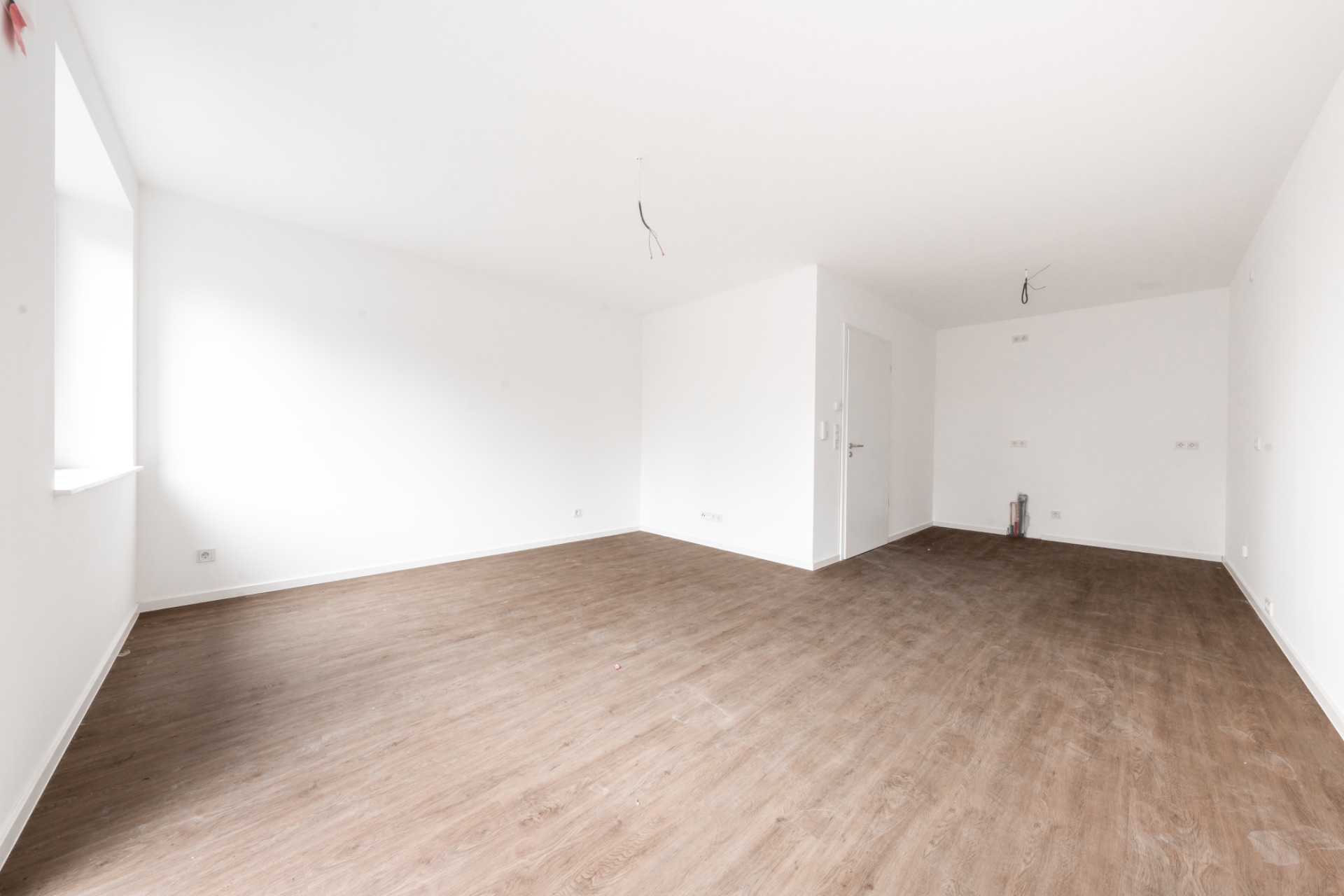 Leerer Raum mit weiß gestrichenen Wänden und braunem Holzboden, Fenster auf der linken Seite.
