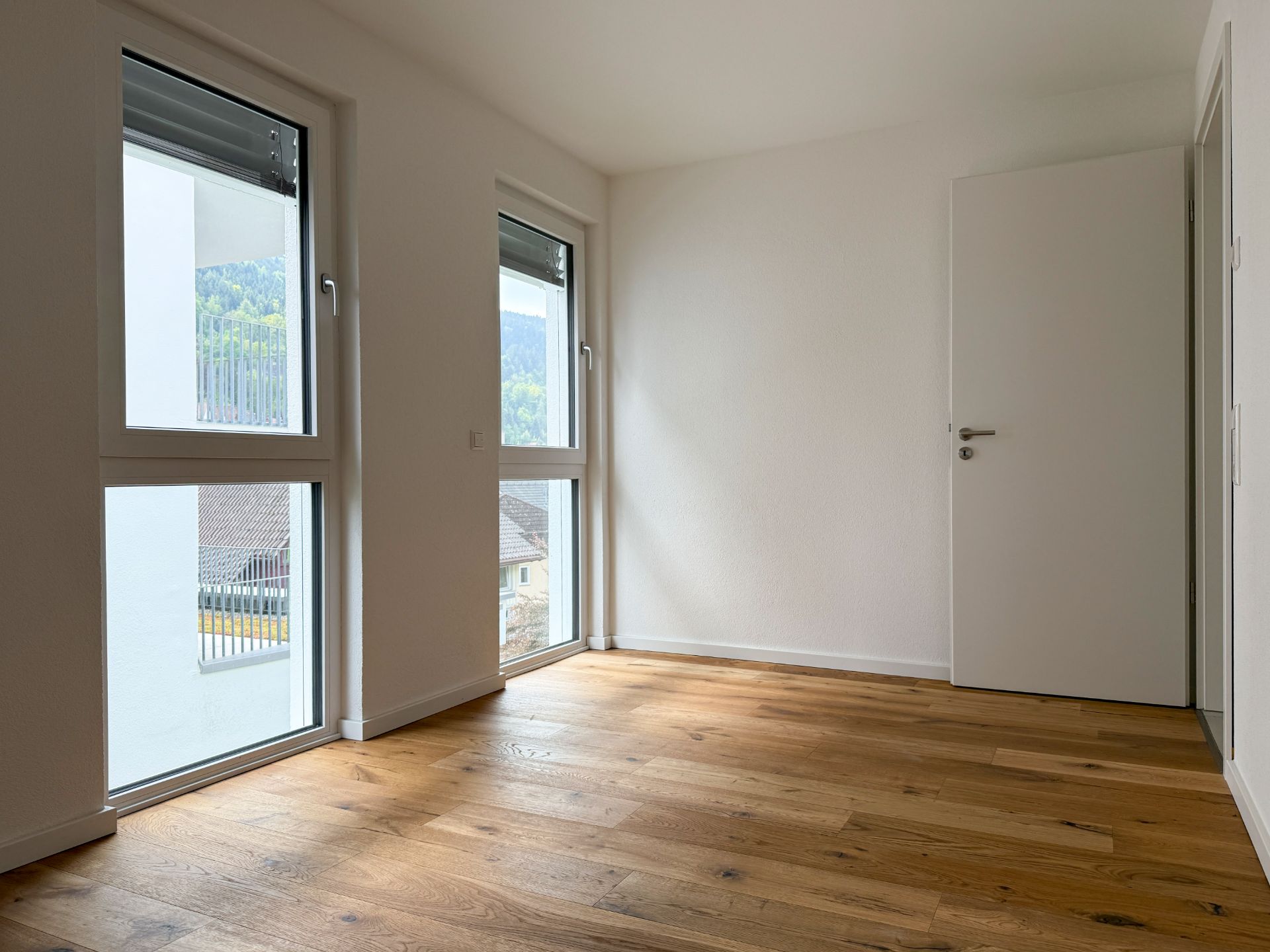 Helles, leeres Zimmer mit Holzfußboden, zwei großen Fenstern und weißer Tür.