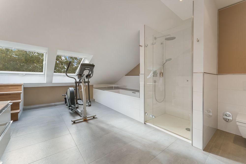 Modernes Badezimmer mit Dusche, Badewanne, Fitnessgerät und zwei Fenstern unter Dachschräge.