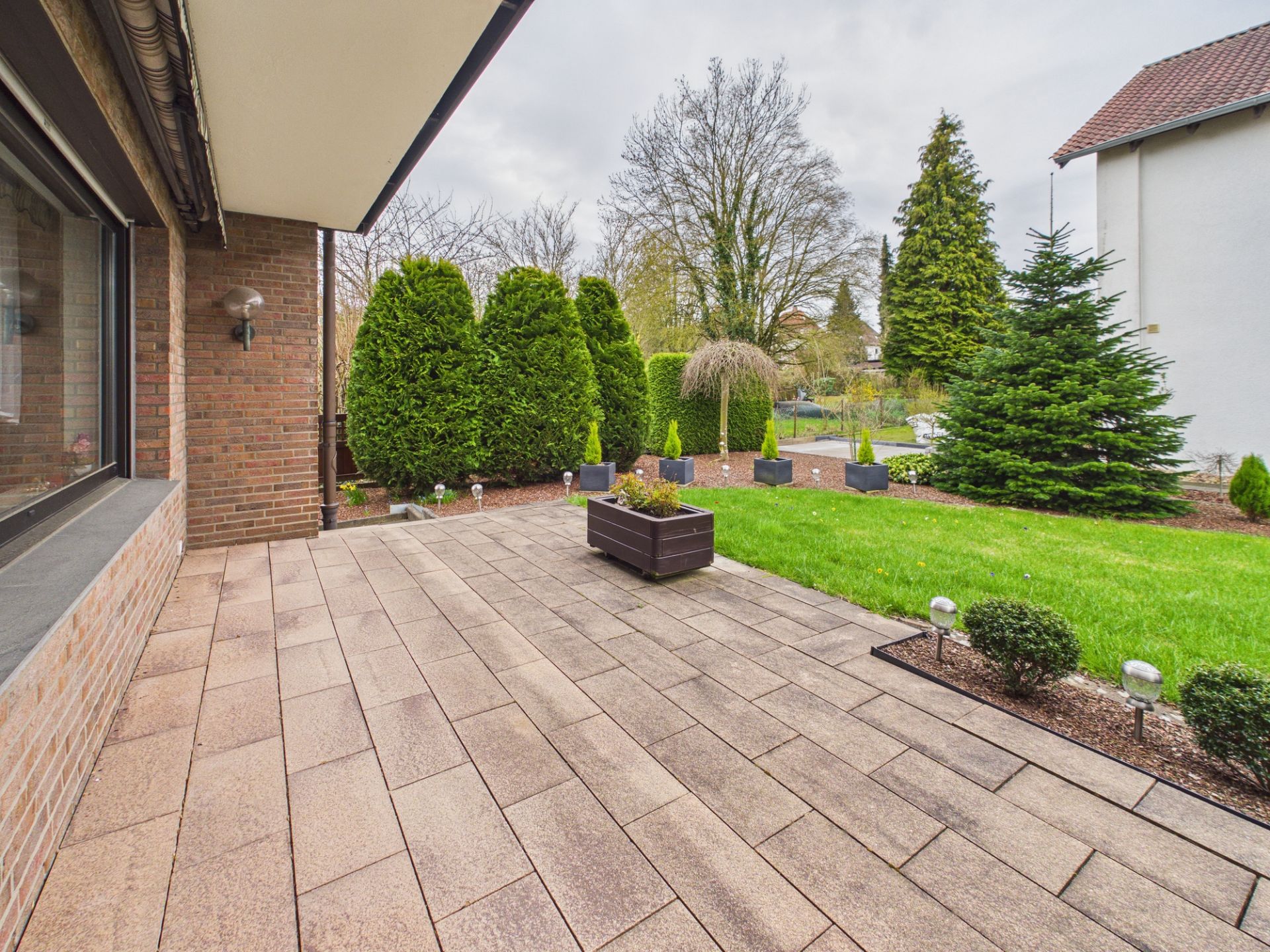 Gepflasterte Terrasse mit Pflanzkübel, daneben Rasen und verschiedene Sträucher im Garten.