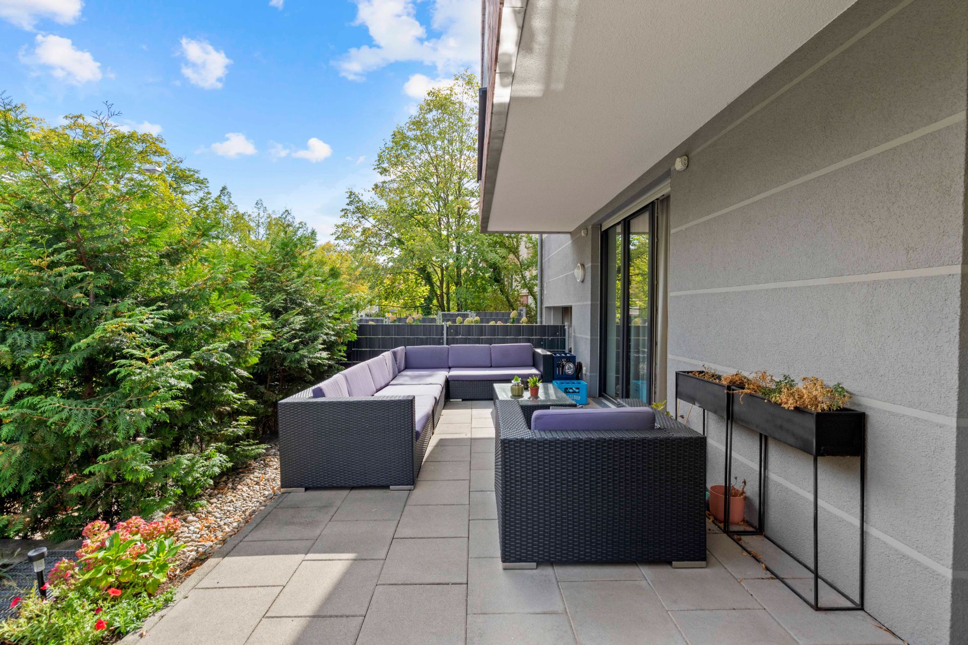 Moderne Terrasse mit Sitzgarnitur, Pflanzen und Fliesenboden, umgeben von grünen Büschen.