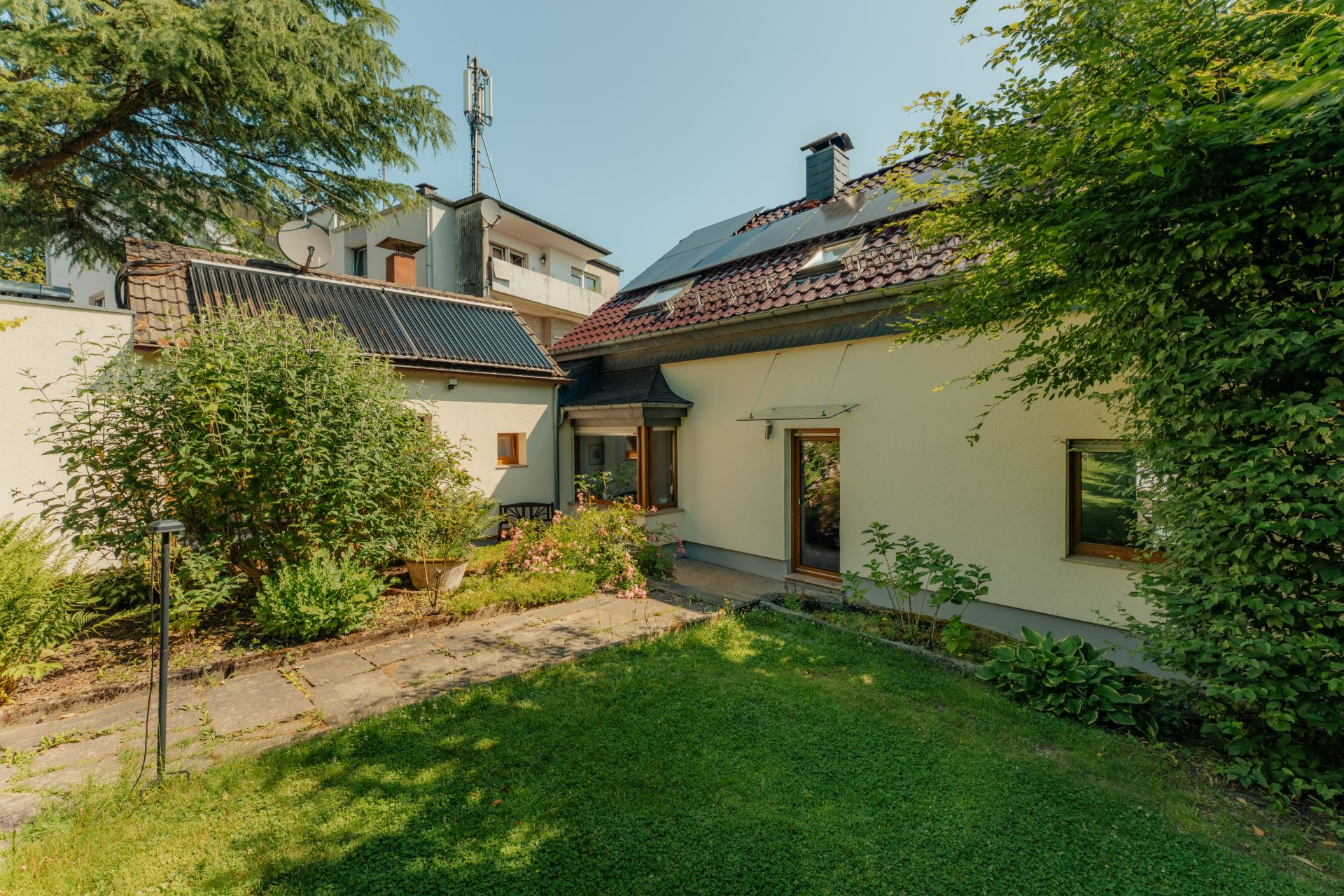 Haus mit Garten, Grünflächen und Sträuchern an einem sonnigen Tag.